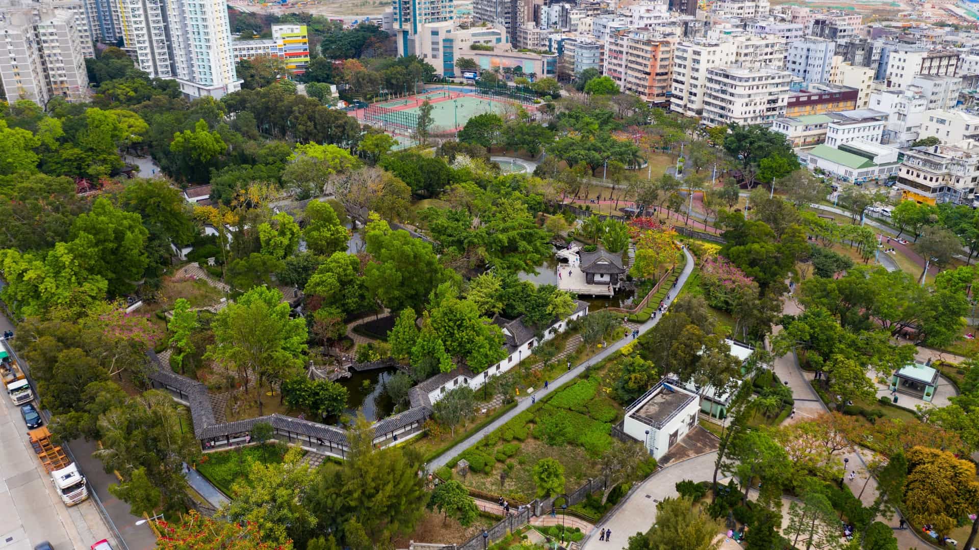 A high angle, top down view of Kowloon Walled City Park in Hong Kong. The image shows a serene, classical Chinese garden with a large pond at its center. The park features lush greenery, traditional Chinese pavilions with curved roofs, and intricate pathways. The surrounding urban landscape with modern buildings is visible in the background, contrasting with the tranquility of the park.