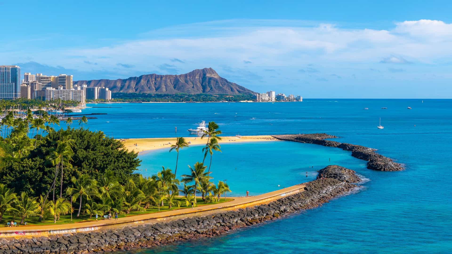 An aerial view of Waikiki Beach in Honolulu, Oahu, Hawaii. The image shows the curved white sand beach and the turquoise waters of the Pacific Ocean. High rise hotels and resorts line the coastline, with a clear view of the city skyline. In the distance, the iconic Diamond Head crater is visible, providing a backdrop of lush green hills.