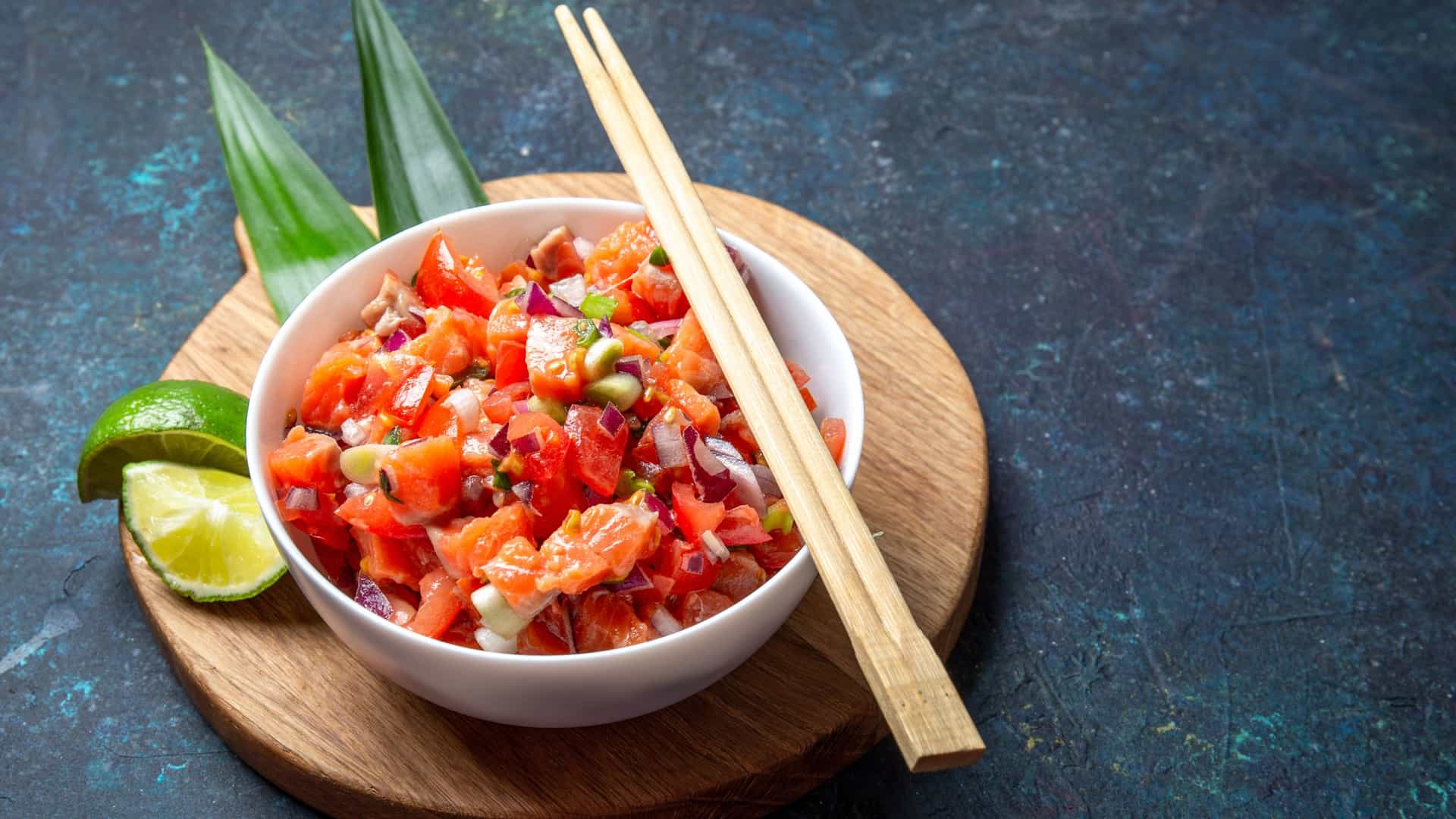 A close up, top down view of a serving of Lomi Salmon, a traditional Hawaiian raw salad. The dish consists of small, cubed pieces of bright pink salted salmon mixed with diced red tomatoes, white onions, and finely chopped green onions. The vibrant colors of the ingredients are clearly visible, and the salad is served in a dark colored bowl, which provides a strong contrast.