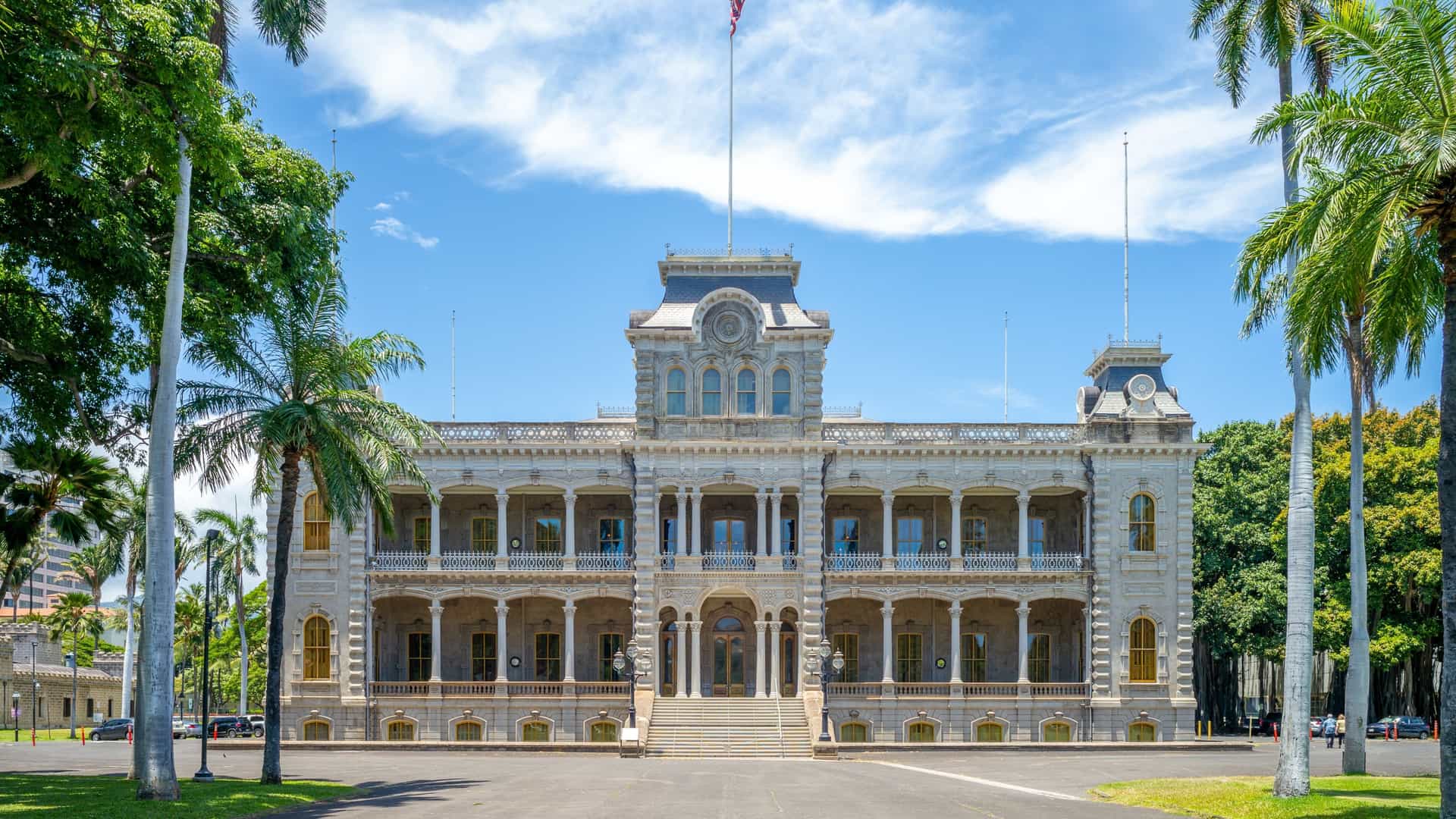 An exterior view of the ʻIolani Palace in Honolulu, Hawaii. The building, constructed in the American Florentine style, is a two story structure with four corner towers and two center towers. The palace is made of brick with concrete facing and features double lanais (verandas) on the first and second floors. The image captures the historical landmark against a backdrop of a clear sky, with manicured green lawns and palm trees surrounding the building. The Iolani Coronation Pavilion is also visible on the palace grounds.