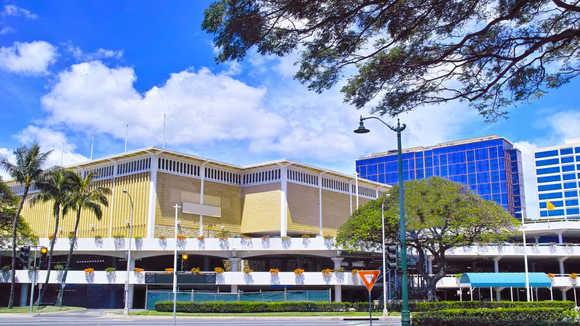 An exterior view of the multi-level, open-air Ala Moana Center in Honolulu, Hawaii, the world's largest open-air shopping mall. The photo captures the modern architecture of the center, a common shopping destination for residents and tourists on the island of Oahu.