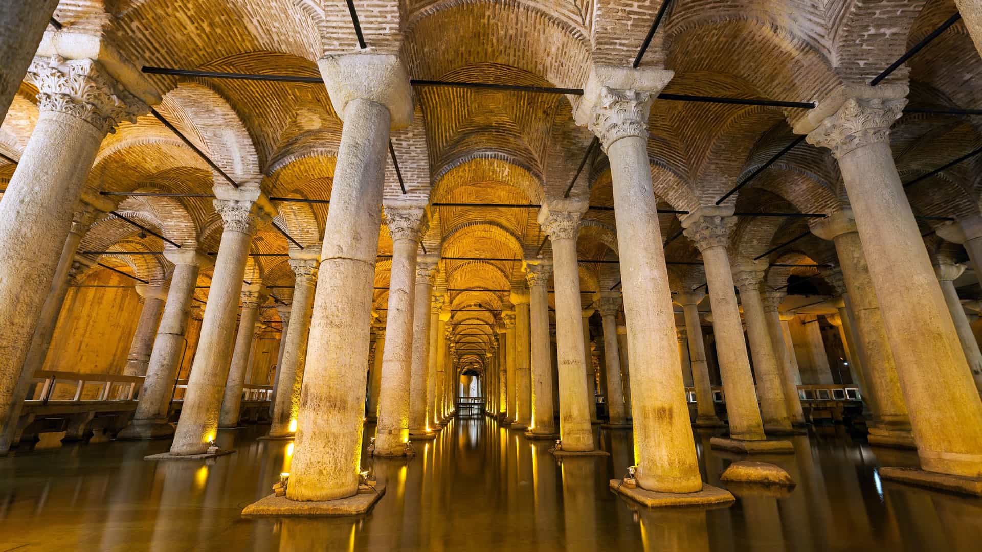 A view of the Basilica Cistern, a historic underground water reservoir in Istanbul, Turkey. The image shows the dimly lit, vast chamber filled with a shallow layer of water. The vaulted ceiling is supported by a grid of 336 towering marble columns that are illuminated from below, creating a mesmerizing and reflective visual effect.