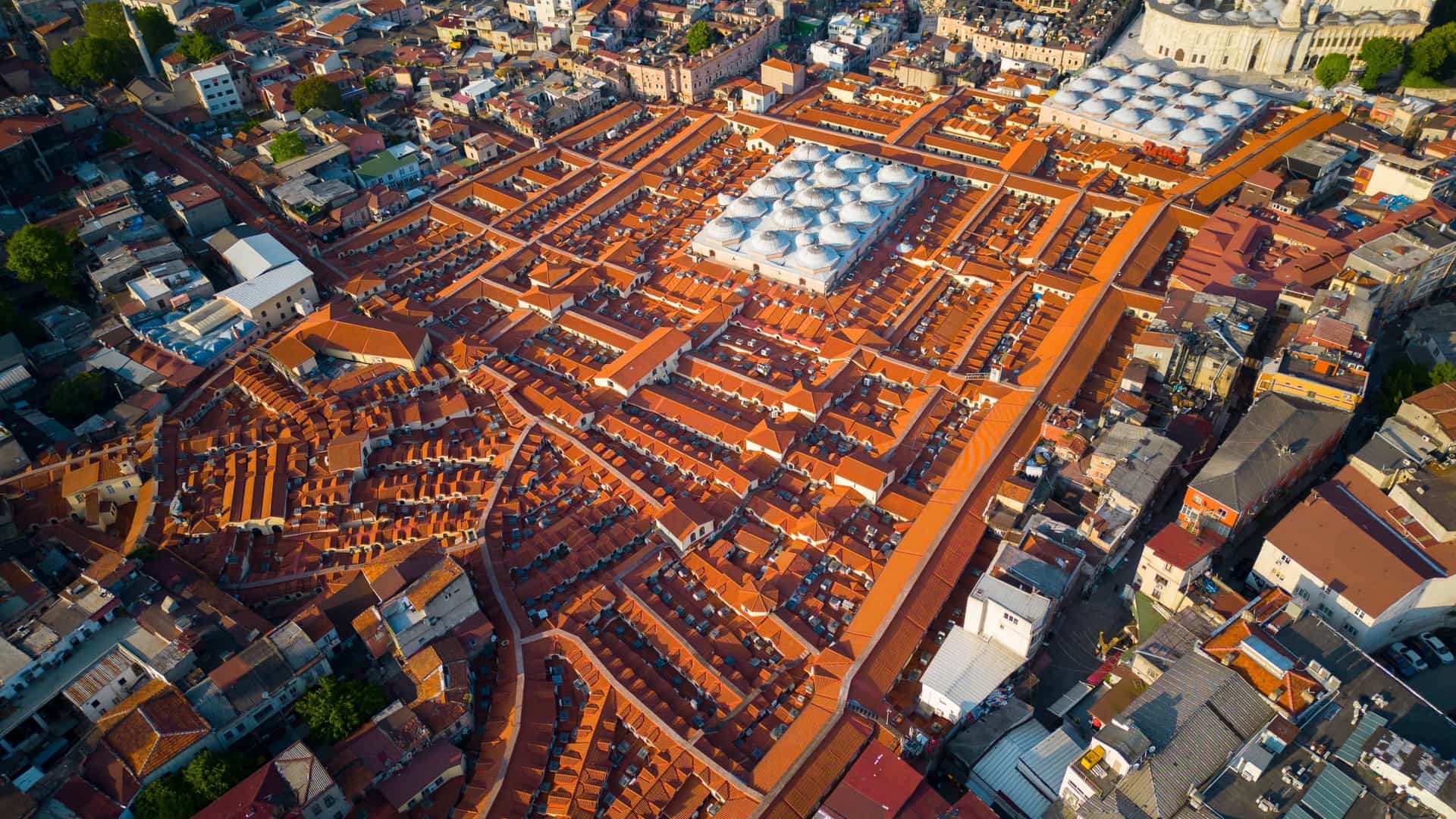 An aerial view of the Grand Bazaar in Istanbul, Turkey. The image shows the vast and dense network of domed rooftops that cover the historic market. The domes, arranged closely together, form a textured and unique pattern, surrounded by other buildings and streets of the Fatih district.
