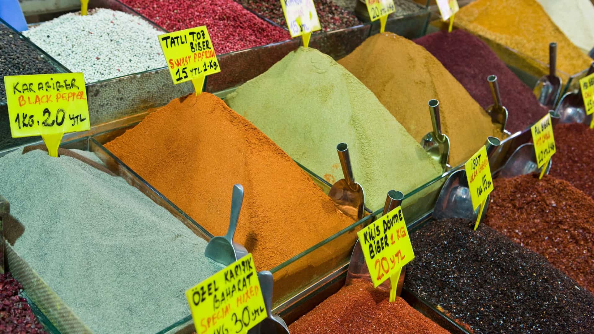 A vibrant photo of a vendor's stall at the Spice Bazaar in Istanbul, Turkey. The image is filled with large, cone shaped mounds of colorful spices and herbs, including various shades of red, yellow, and green. The spices are displayed in open containers, creating a rich texture and a feast for the eyes, typical of the bustling market.