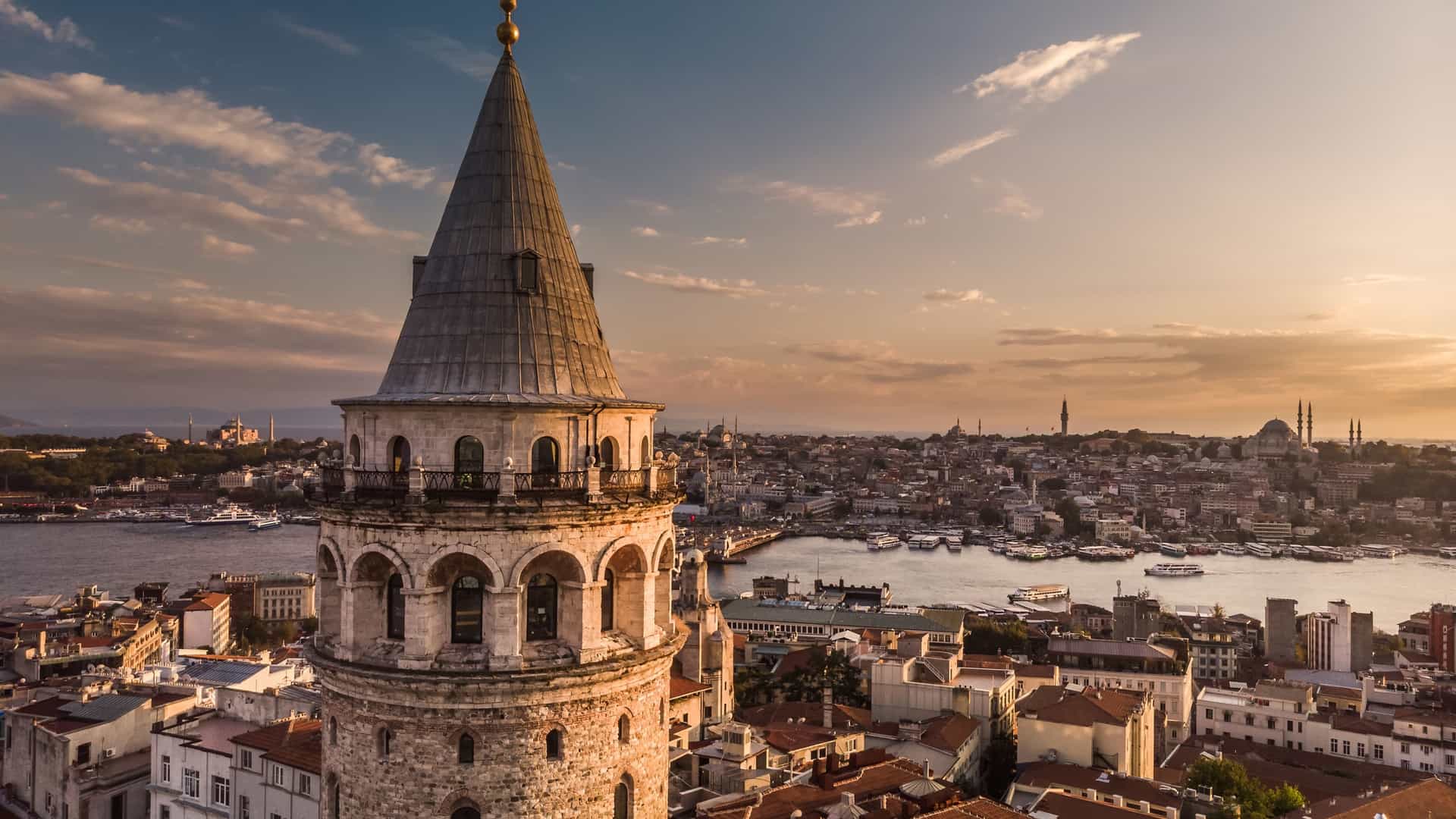 An aerial, evening shot of the Galata Tower in Istanbul, Turkey. The medieval stone tower is brightly lit, standing prominently among the city's rooftops. The image captures the cityscape at dusk during the "blue hour," with the lights of the buildings beginning to glow against the darkening sky and a body of water, likely the Golden Horn or Bosphorus, visible in the background.