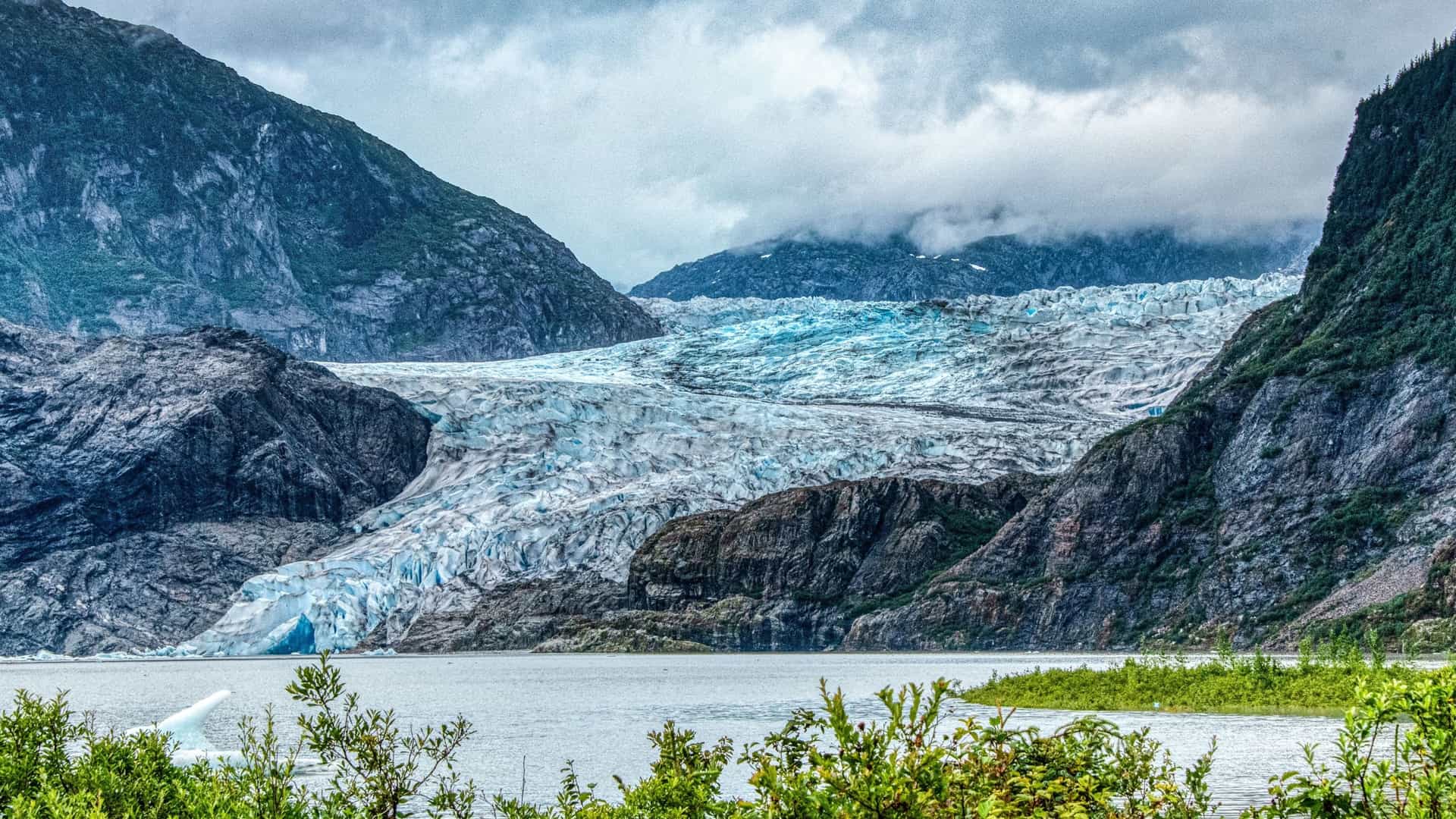 A scenic view of the Mendenhall Glacier in Juneau, Alaska. The glacier, a massive river of ice, flows down from the mountains and ends at a lake. The scene is surrounded by rugged, forested mountains, and the sky is overcast.