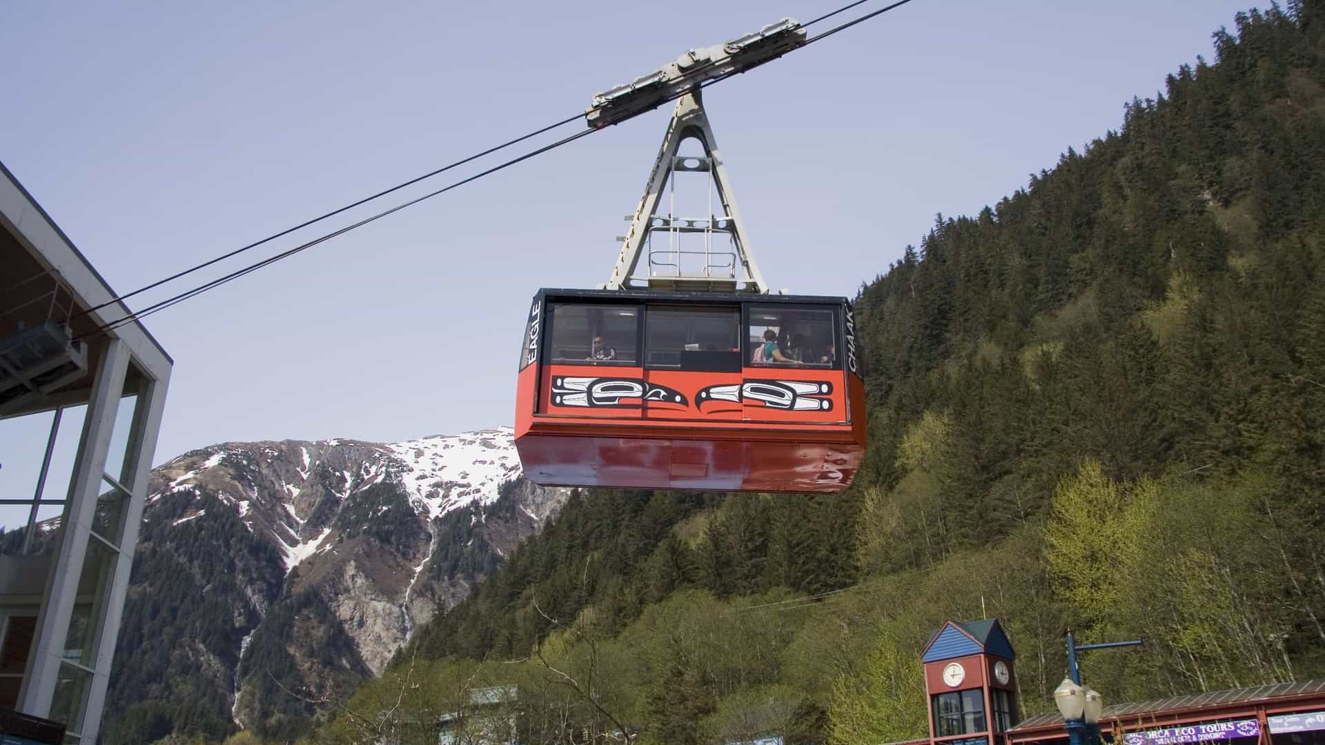 A scenic photograph of the red tram car of the Mount Roberts Tramway in Juneau, Alaska, ascending the steep, forested mountainside. The tram is carrying riders to the summit, offering a view of the mountains and the surrounding landscape.