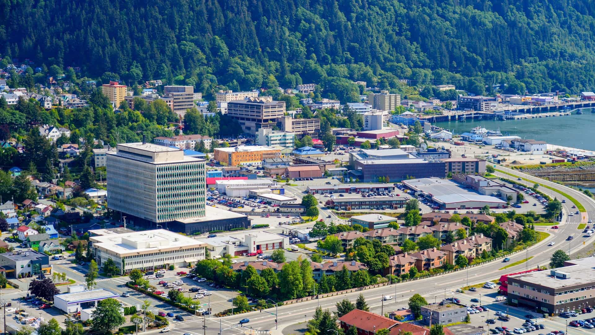 An aerial photograph providing a panoramic view of downtown Juneau, the state capital of Alaska, nestled between the waterfront of the Gastineau Channel and the base of steep, forested mountains. The image shows the compact grid of the city, including its docks, urban buildings, and surrounding wilderness.