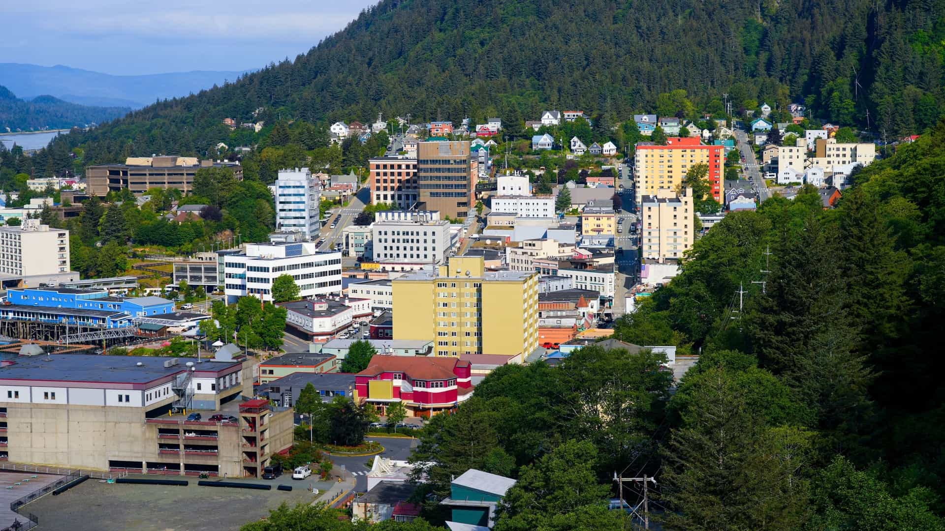 An aerial view of downtown Juneau, Alaska, showing the urban layout of the state capital nestled against the steep, forested mountains. The photograph includes the commercial waterfront area, where structures like the famous Red Dog Saloon are located near the docks and modern buildings.