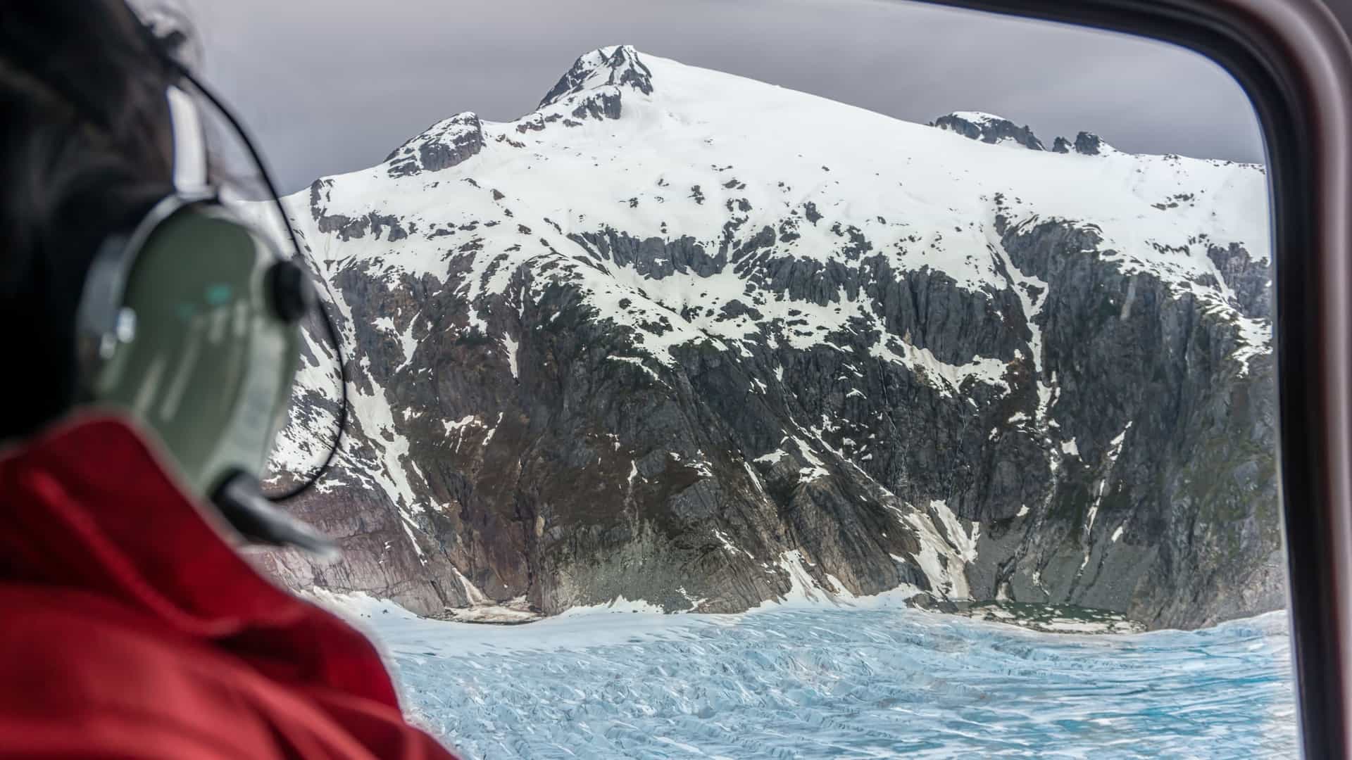 An in-flight view from the passenger window of a helicopter, looking out onto a vast, crevassed glacier and snow-capped mountain peaks. The scene captures the extreme, icy terrain of a glacier, often associated with Alaskan sightseeing tours.
