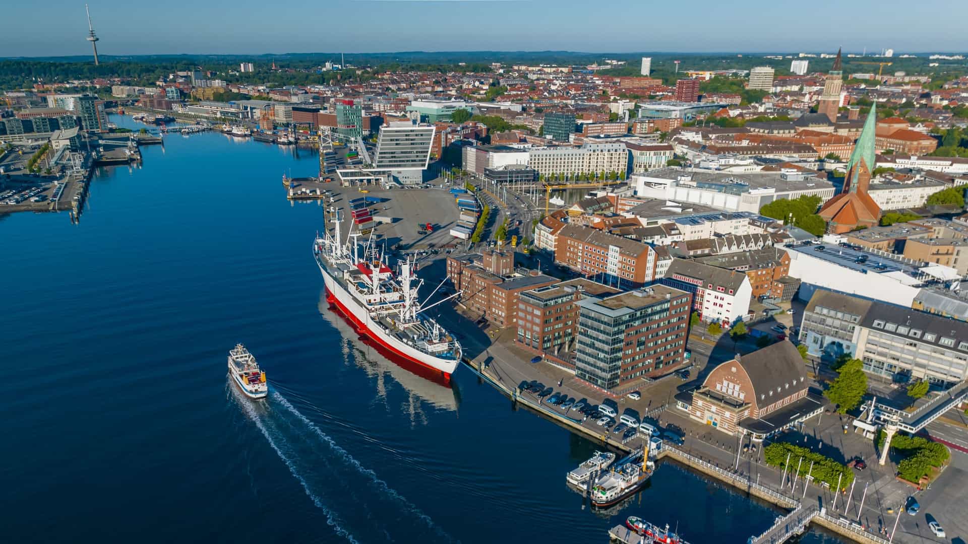 An aerial view of the Port of Kiel in Schleswig-Holstein, Germany, capturing the inner harbor and its various docks. The historic building housing the Kiel Maritime Museum (formerly the fish auction hall) is prominently visible along the waterfront, alongside its collection of historic museum ships moored at the adjacent pier.