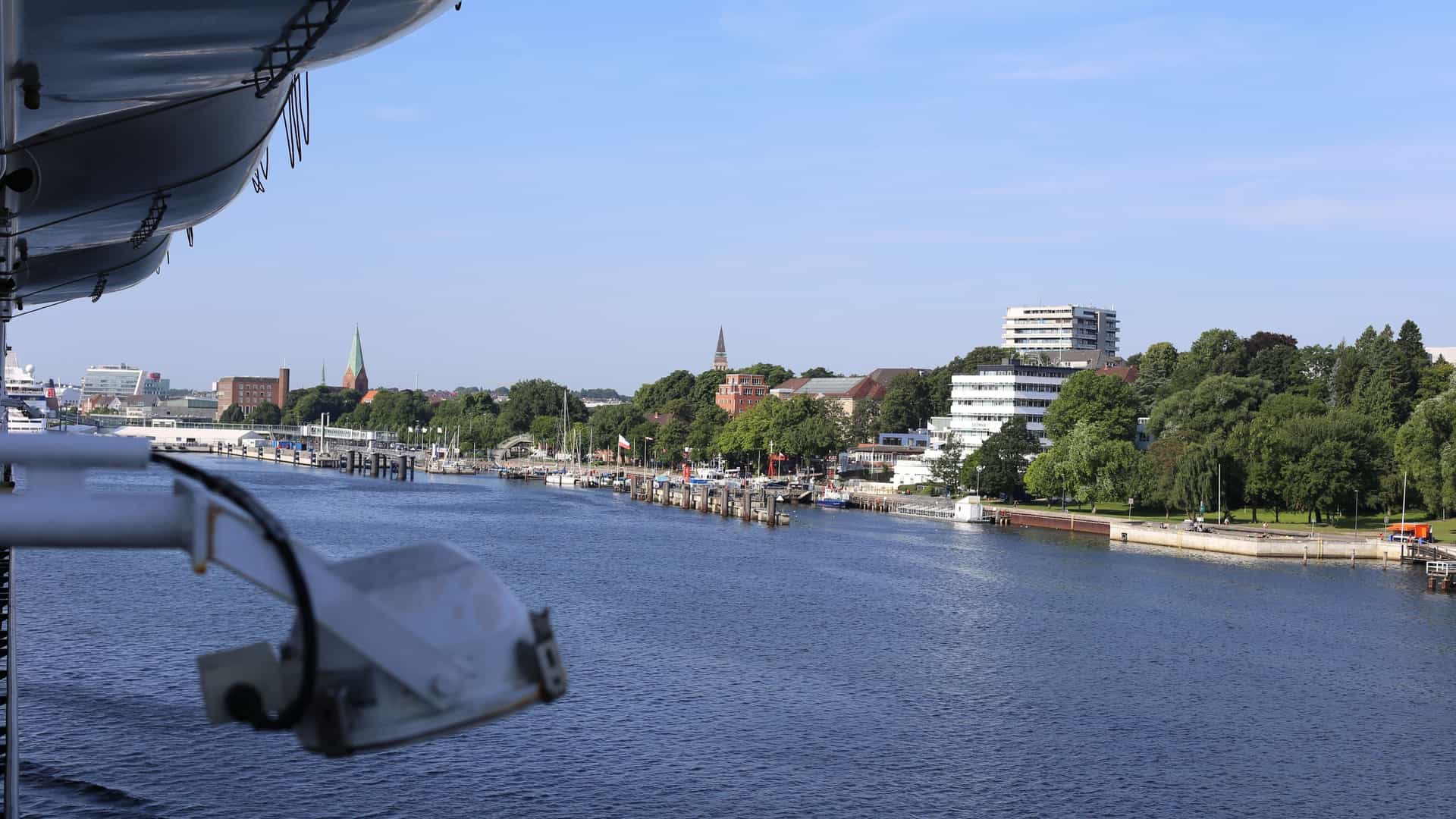 A view from a ship or ferry looking toward the shore, capturing the Kiellinie, the popular waterfront promenade in Kiel, Germany. The image shows a long stretch of the shore lined with boat docks, green trees, and the city's skyline, including the spires of historic churches, under a blue sky.
