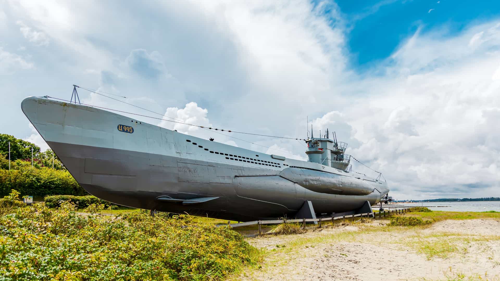 A large, full-view exterior shot of the German Type VIIC/41 U-Boat U-995, a historic World War II submarine, resting permanently on the beach in Laboe, Germany, near the Laboe Naval Memorial. The massive, black-painted submarine, preserved as a technical museum, is visible against the sand, with its conning tower and hull dominating the foreground.