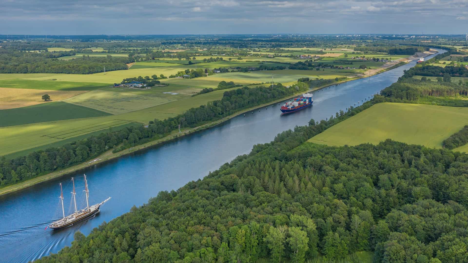 An aerial shot of the Kiel Canal in Germany, showing a large container ship and a traditional sailing vessel. The canal, which connects the Baltic and North Seas, is bordered by green fields. The image captures the contrast between modern commercial shipping and historical sailing on this major waterway.