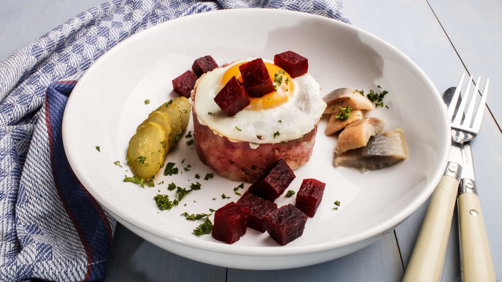 A close up of a traditional North German dish called Labskaus. It is a reddish pink mash made of potatoes, corned beef, and beetroot. The dish is served with a fried egg on top, a pickle,