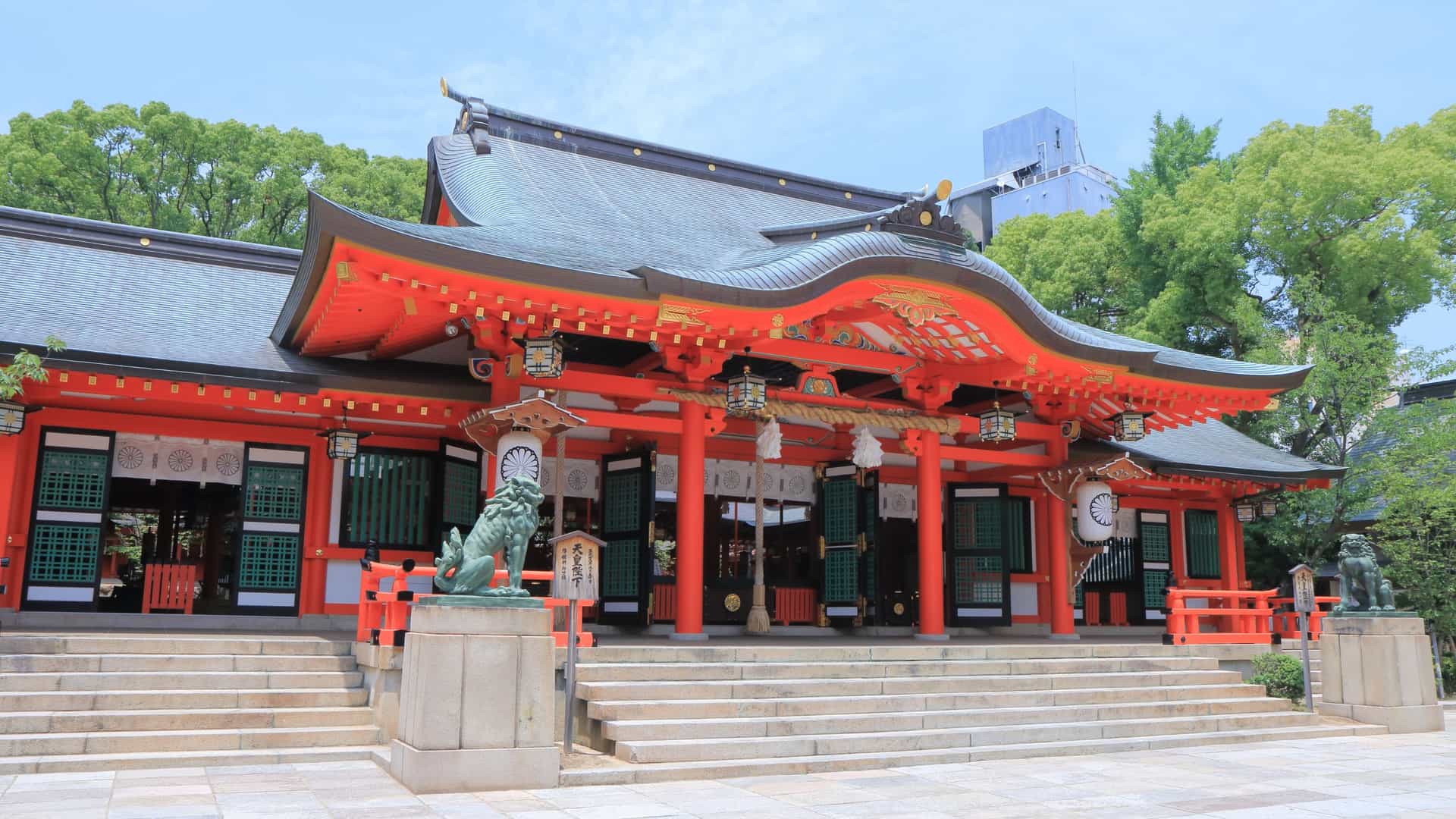 A scenic view of Ikuta Shrine in Kobe, Japan. The image shows the prominent red wooden tower gate, or torii, surrounded by lush green trees. The shrine is located in the heart of the city, providing a serene and traditional contrast to the modern urban surroundings.
