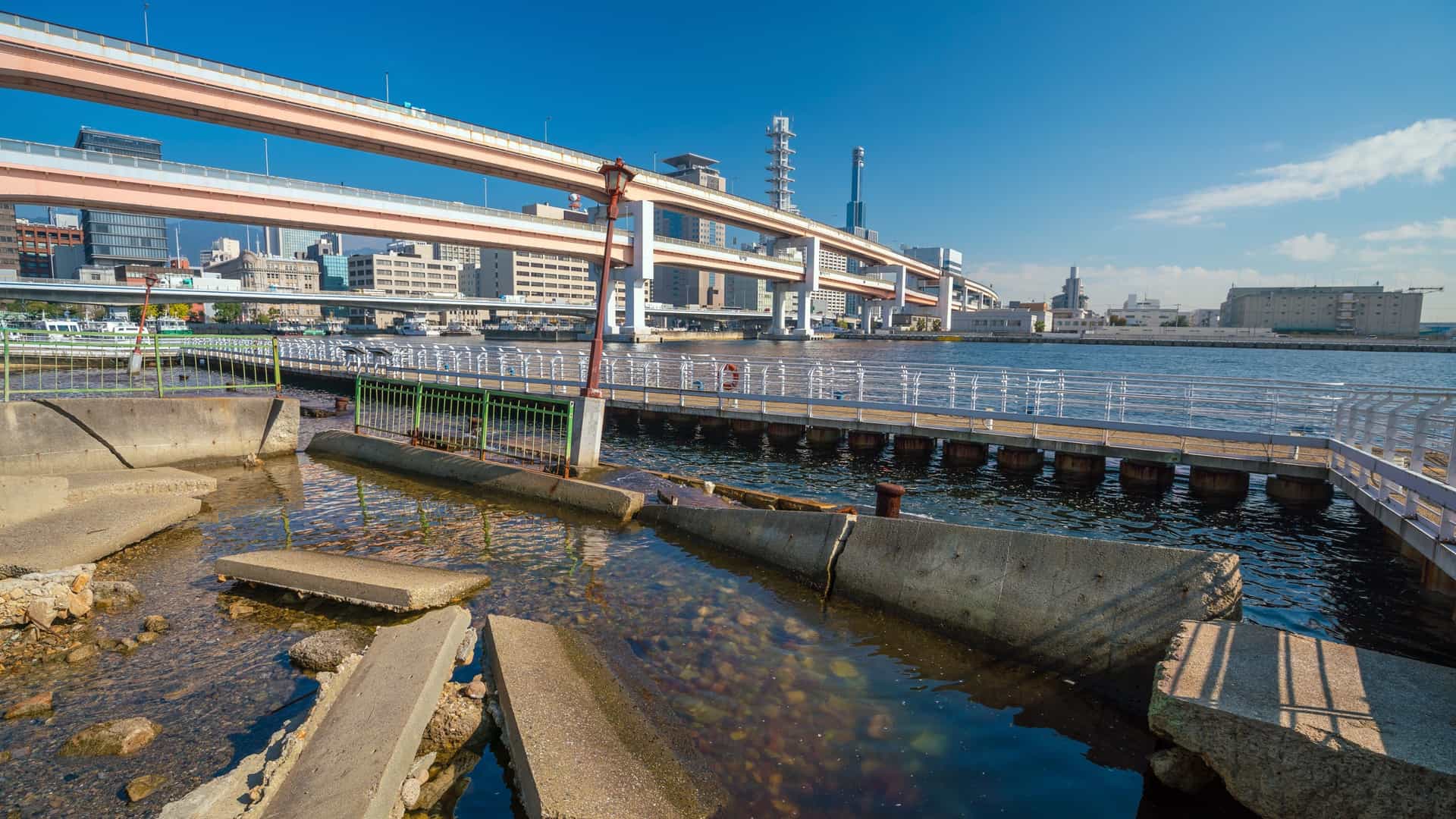 A view of the Kobe Earthquake Memorial Park in Kobe, Japan, a memorial site that preserves a portion of the collapsed quay wall from the 1995 Great Hanshin Awaji Earthquake. The image shows the buckled and twisted concrete pier and ground, a stark reminder of the earthquake's destructive power.