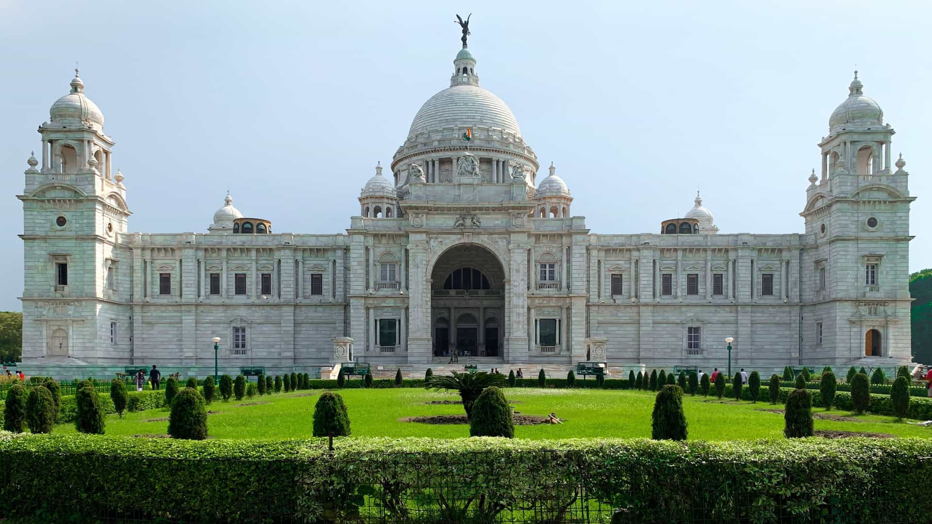 The towering, brilliant white marble Victoria Memorial in Kolkata, India, dominates the sunny, wide-angle view, surrounded by green lawns and gardens, a stunning symbol of colonial architecture and history.