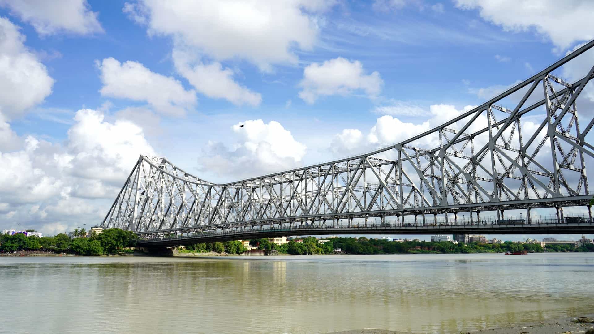 A wide-angle view of the iconic Howrah Bridge, a massive steel truss bridge, spanning the Hooghly River (Ganga) in Kolkata, India. The bridge's industrial steel structure dominates the frame, set against a hazy sky, with boats and water traffic visible on the river below.