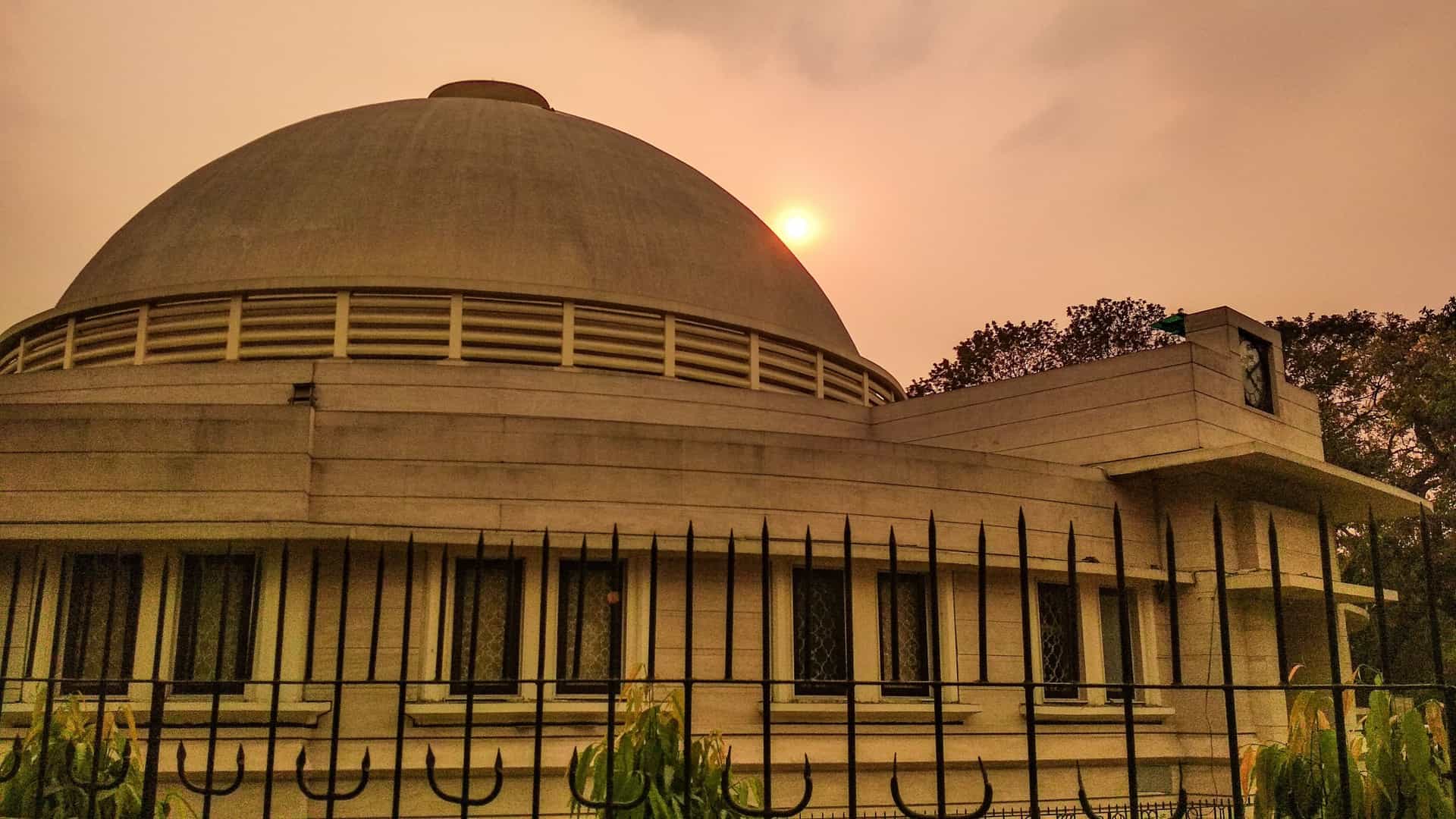 The distinctive, circular, pale brown M.P. Birla Planetarium in Kolkata, India, architecturally resembles a Buddhist stupa. The prominent, dome-shaped building is surrounded by well-kept green lawns and trees, highlighting its stature as a famous city landmark.