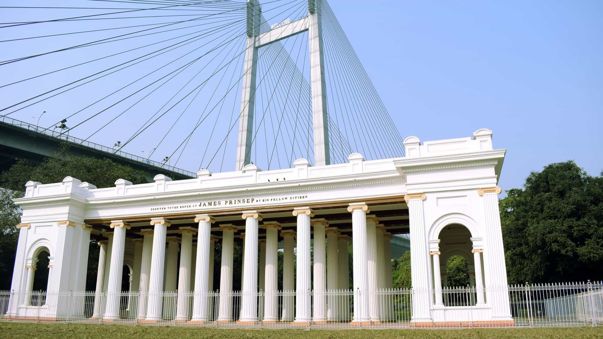 The neoclassical white Palladian James Prinsep Monument stands on the bank of the Hooghly River in Kolkata. The structure's prominent columns frame the modern Vidyasagar Setu (Second Hooghly Bridge) in the distance, contrasting colonial heritage with contemporary infrastructure.