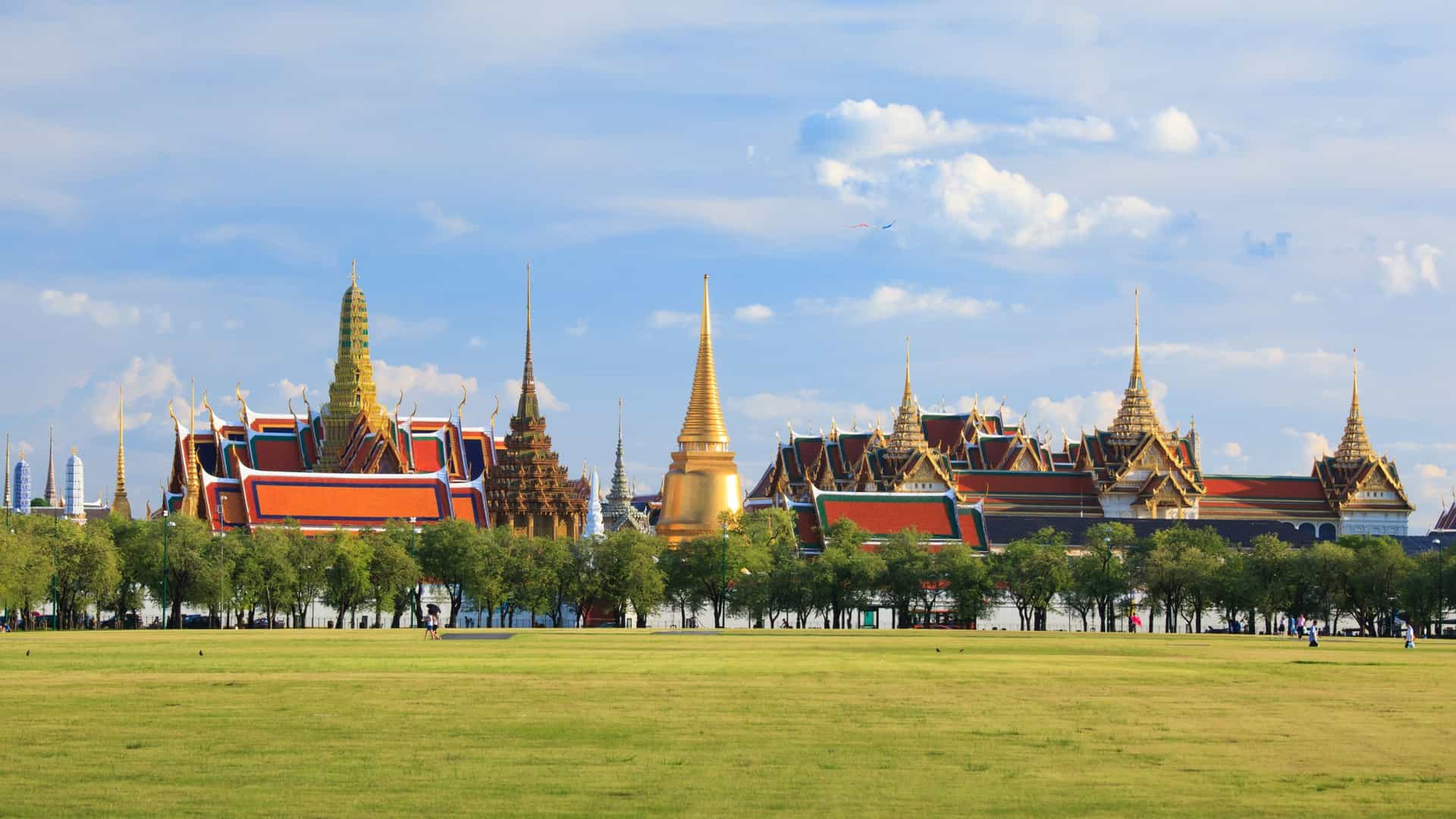 The ornately decorated buildings of Wat Phra Kaew (Temple of the Emerald Buddha) are shown under a blue sky, with golden stupas, colorful spires, and detailed rooflines. The complex is a famous landmark within the Grand Palace in Bangkok, Thailand.
