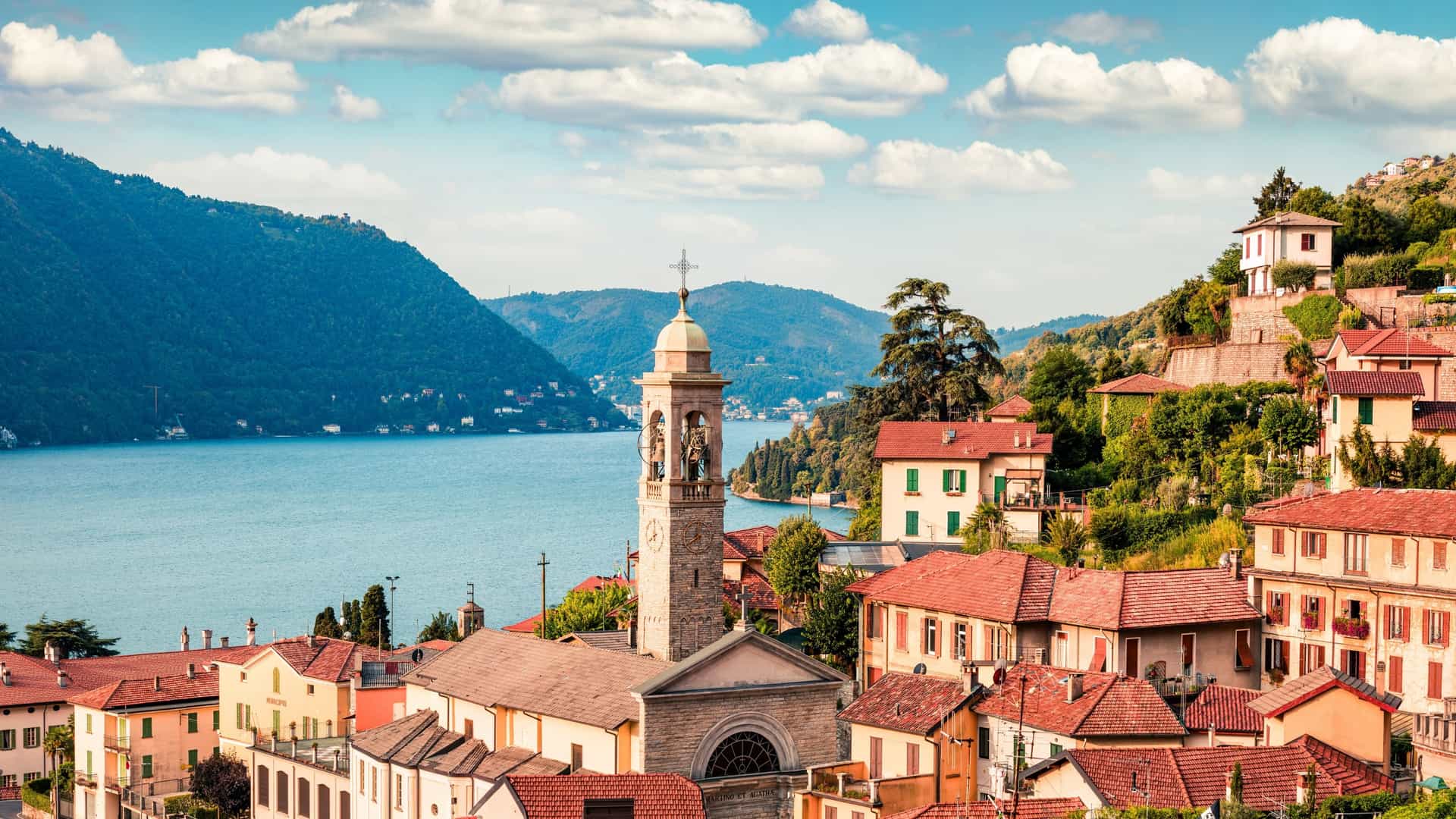 A breathtaking view of Lake Como in Italy on a sunny day. The clear blue water of the lake is surrounded by lush green mountains and charming villas with red roofs. A boat is visible on the water, leaving a white wake behind it. The sky is a clear blue with a few fluffy white clouds.