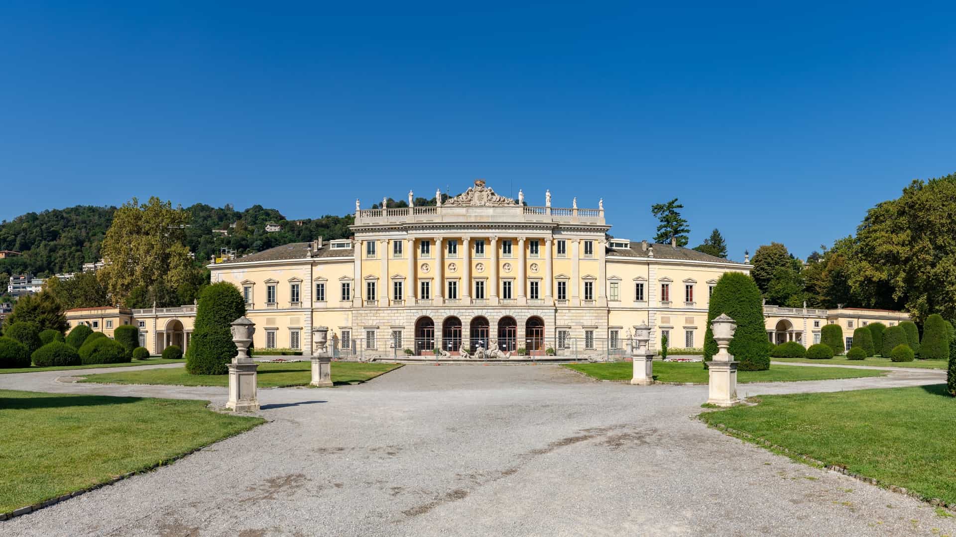 A view of Villa Olmo, a grand neoclassical building in Como, Italy, situated on the shores of Lake Como. The villa's ornate, symmetrical facade with columns is visible, surrounded by manicured green lawns and trees in its extensive garden.