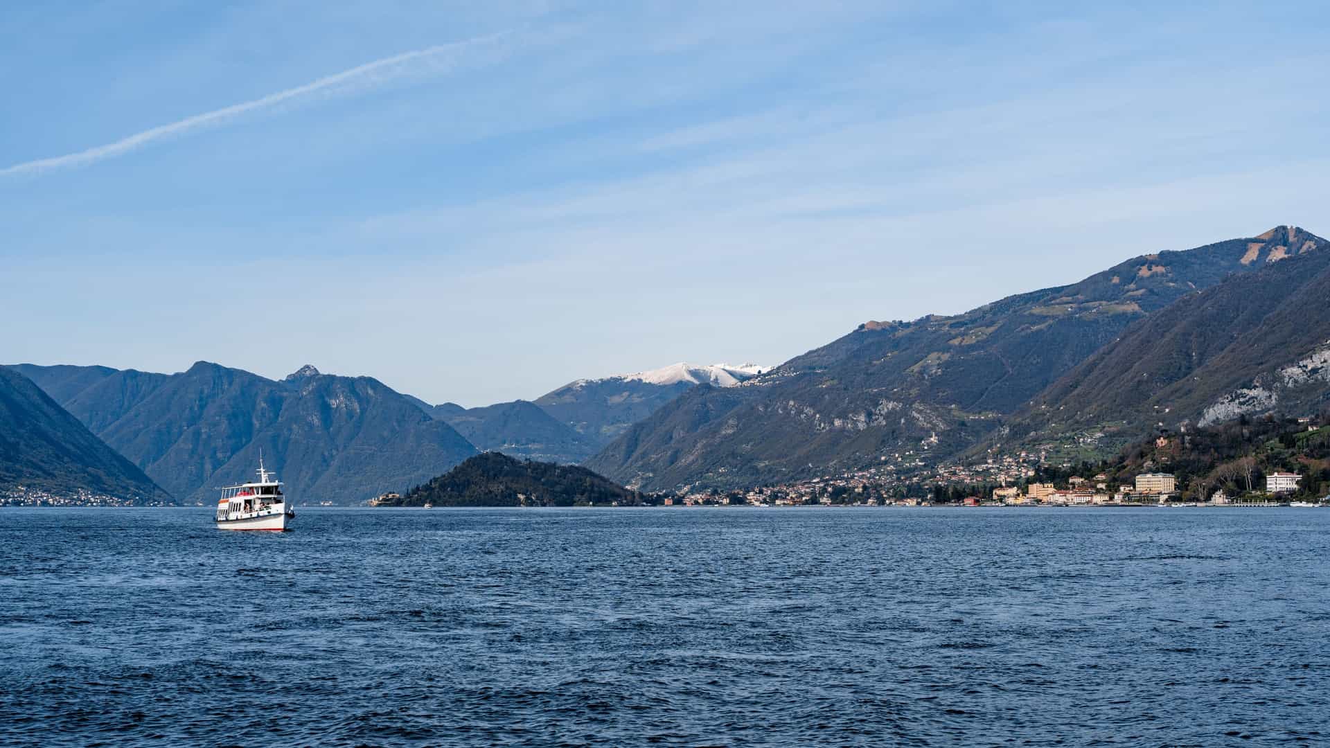 A ferry is sailing on the calm waters of Lake Como in Italy, with majestic mountains and a clear sky in the background. The boat is leaving a white wake behind it.