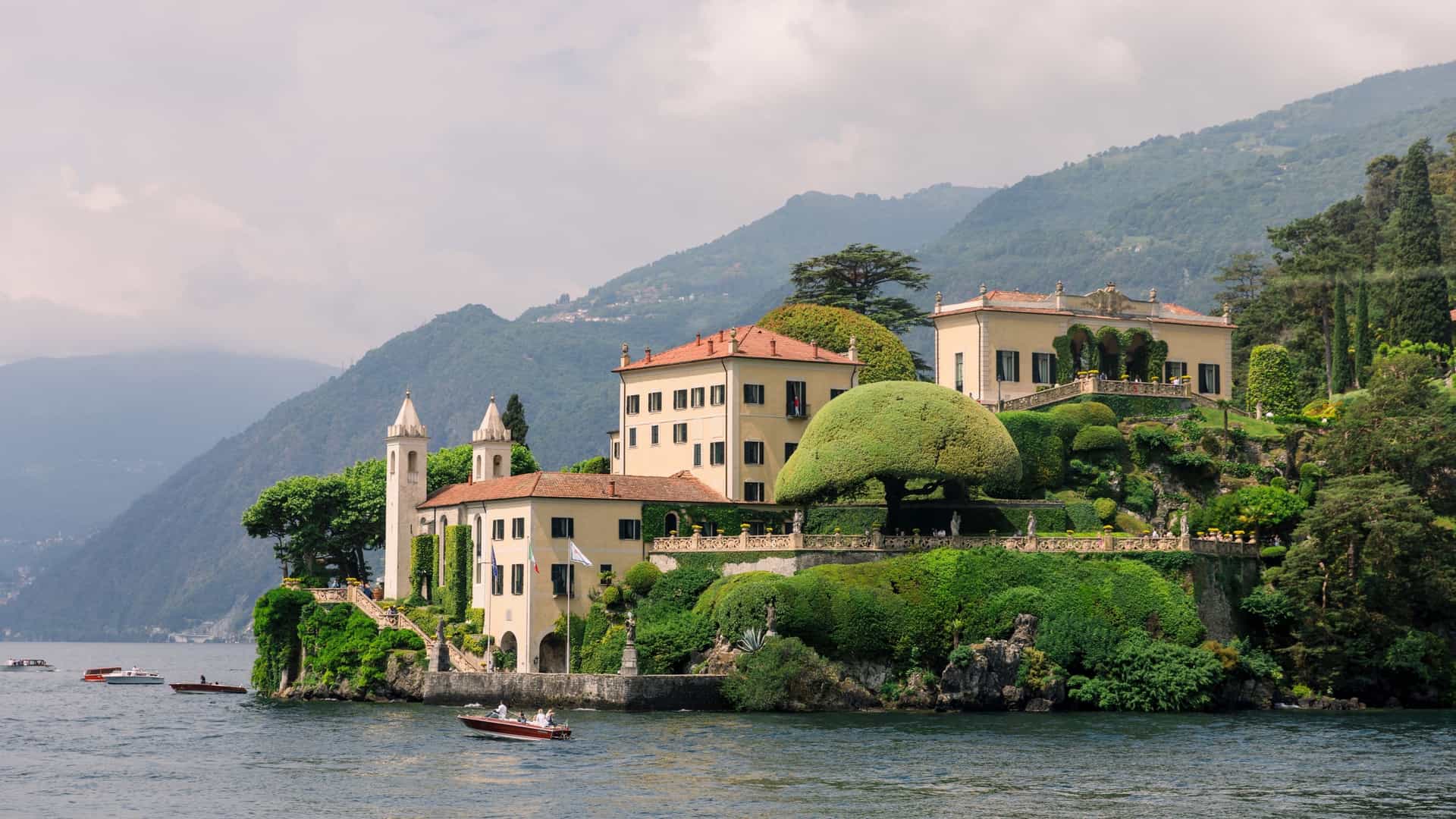 An elevated view of Villa del Balbianello, a terraced villa with an elaborate garden situated on a peninsula on the western shore of Lake Como, Italy. The photo shows the loggia and a lush garden with an umbrella pruned tree, with the calm lake and mountains in the background.