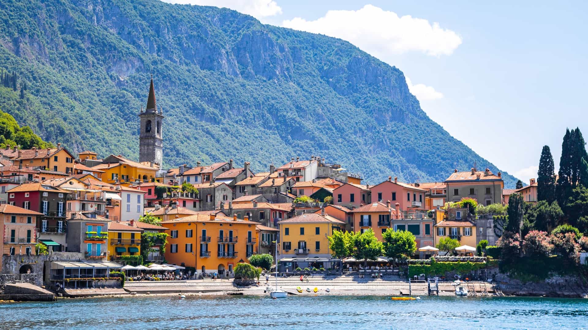 A view looking down a narrow, stepped cobblestone street in the town of Varenna, Italy. The street is lined with colorful old buildings, and in the distance, a glimpse of Lake Como and the mountains beyond is visible.