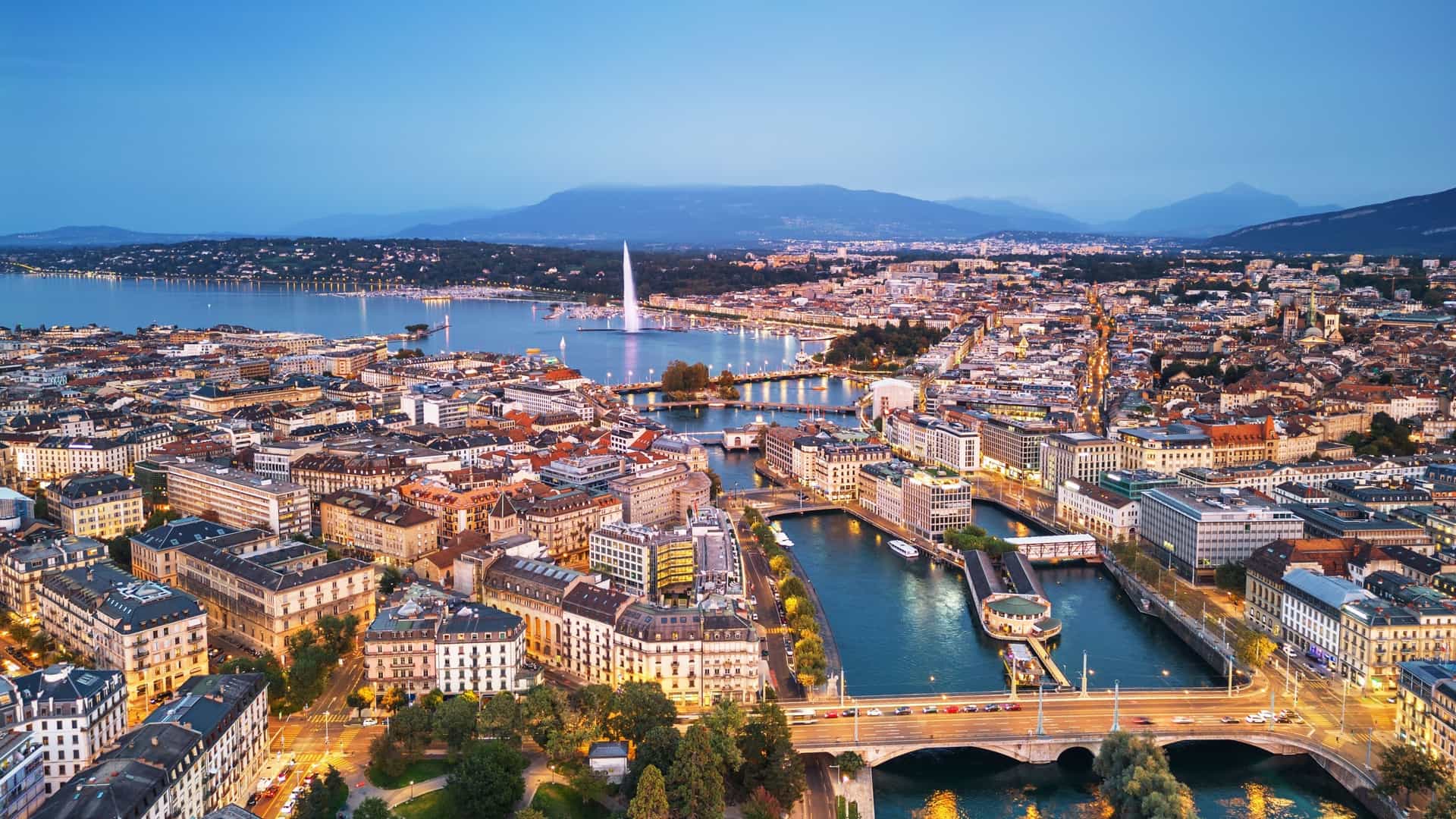 A view of the Geneva, Switzerland, skyline at twilight, with the city's famous Jet d'Eau fountain in the foreground. The powerful water jet is illuminated against the darkening sky, and the city's buildings are visible along the shore of Lake Geneva.