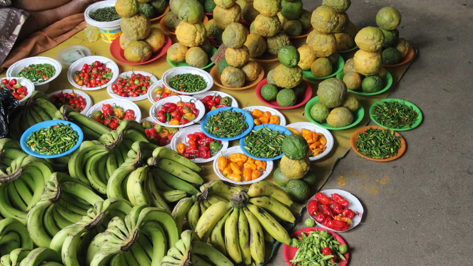 A vibrant display of tropical fruits and vegetables for sale at a stall in the Lautoka Market in Fiji. The photo shows a colorful variety of fresh, local produce, likely including items such as bananas, papayas, chilies, and root vegetables, arranged in baskets or on tables in an indoor market setting.