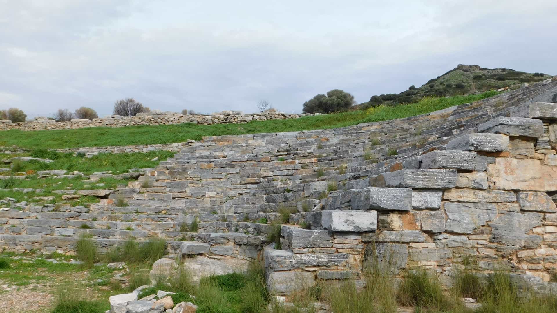 An overall view of the ruins of the Ancient Theater of Thorikos, located on the slope of Velatouri Hill near Lavrio, Attica, Greece. This is the world's oldest known stone theater, built between 525–480 BC, and features an unusual, elongated, elliptical shape with a rectangular orchestra, contrasting with later semi-circular Greek theaters. The stone seating (cavea) is partially preserved, overlooking a valley and the Aegean Sea in the distance.