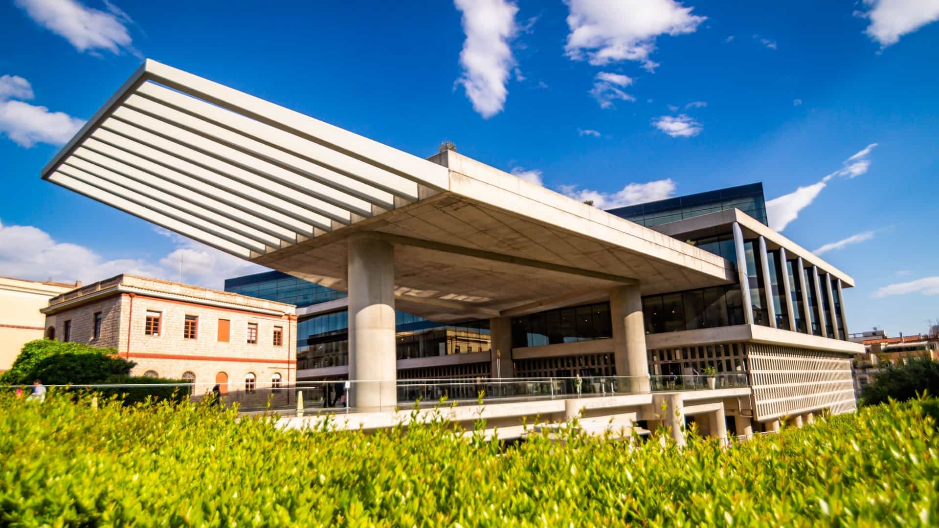 A view of the exterior of the New Acropolis Museum in Athens, Greece. The building, designed by architect Bernard Tschumi, features a striking modern architectural style characterized by simple horizontal lines, massive concrete elements, and extensive use of transparent glass facades. The top-floor Parthenon Gallery, which is rotated to align with the Acropolis, is prominent, allowing a direct visual and contextual connection between the museum and the ancient Parthenon in the background. The base is often seen as being elevated on slender concrete pillars above an archaeological excavation.