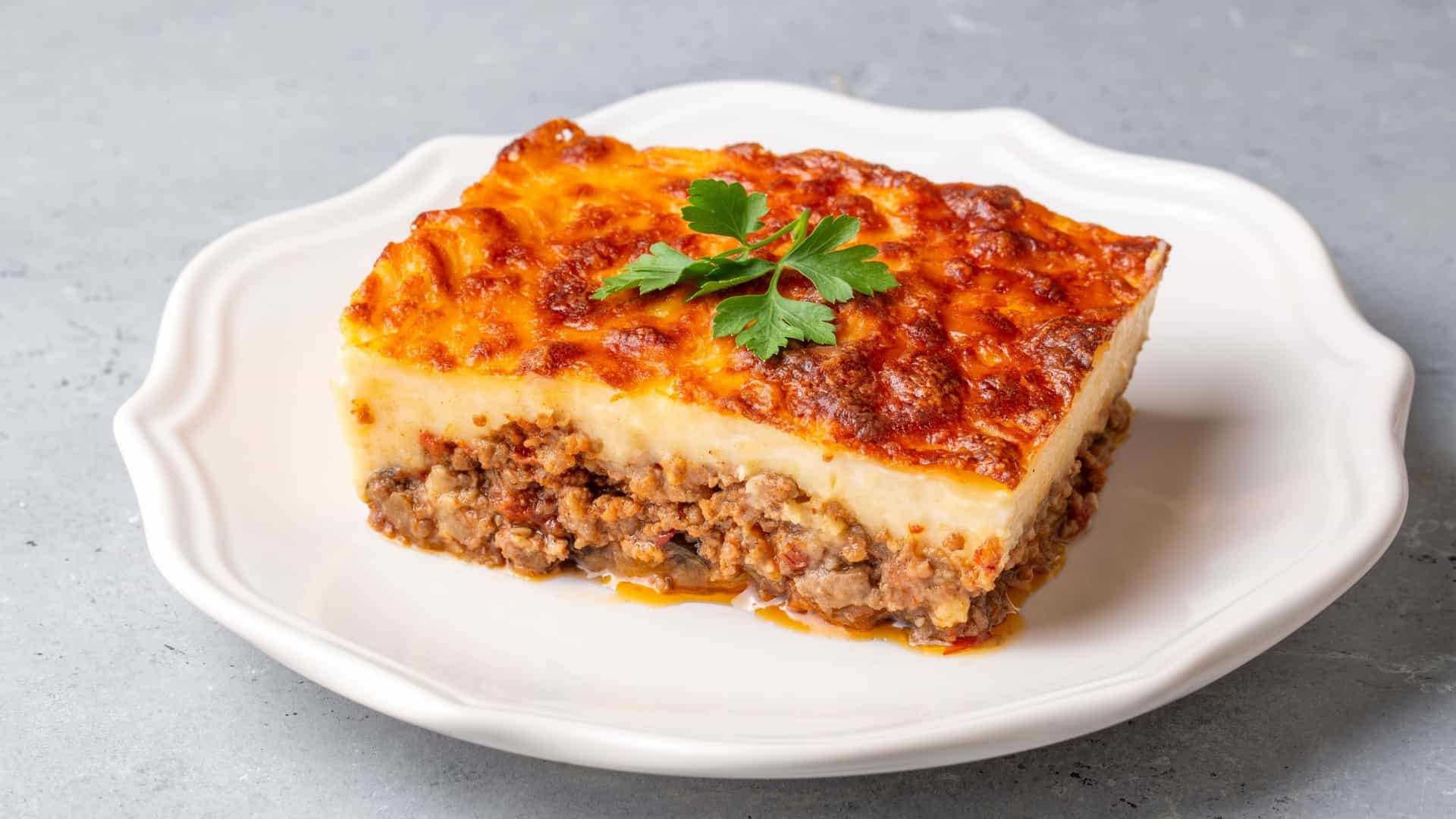 A close-up, appetizing view of a portion of Moussaka, a traditional Greek dish. The casserole is layered, typically featuring slices of eggplant (aubergine), a rich minced meat (ground beef or lamb) and tomato sauce spiced with ingredients like cinnamon and oregano, and is topped with a thick, golden-brown, baked layer of creamy béchamel sauce. The dish is served hot, often on a plate with visible steam or a garnish like a sprig of parsley.