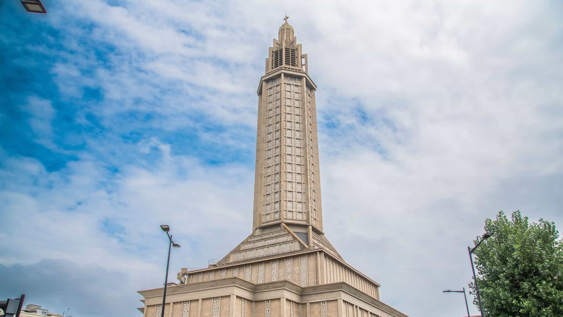 A towering, dramatic exterior view of the Church of Saint Joseph (Église Saint-Joseph) in Le Havre, France. Designed by Auguste Perret, this Modernist and Brutalist concrete structure features a massive, octagonal 107-meter lantern tower. The tower is punctuated by thousands of colored stained-glass windows that serve as a memorial to the civilian victims of World War II, making it an iconic, UNESCO-listed landmark and a lighthouse for the city and port.