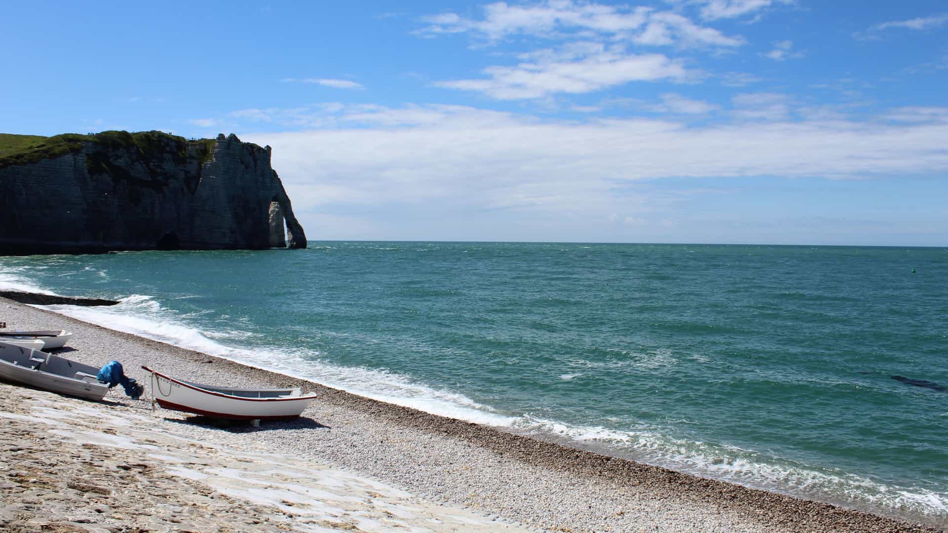 A wide view of the rocky (pebble) beach of Le Havre, France, stretching along the English Channel in the Normandy region. The image captures the extensive shoreline, which consists mainly of large, gray, rounded pebbles (galets). The background features the city's famous modernist seafront architecture—the uniform, post-WWII reinforced concrete buildings—and the long promenade that runs parallel to the water. The beach often shows the contrast between the rough natural stones and the smooth urban lines of the city.