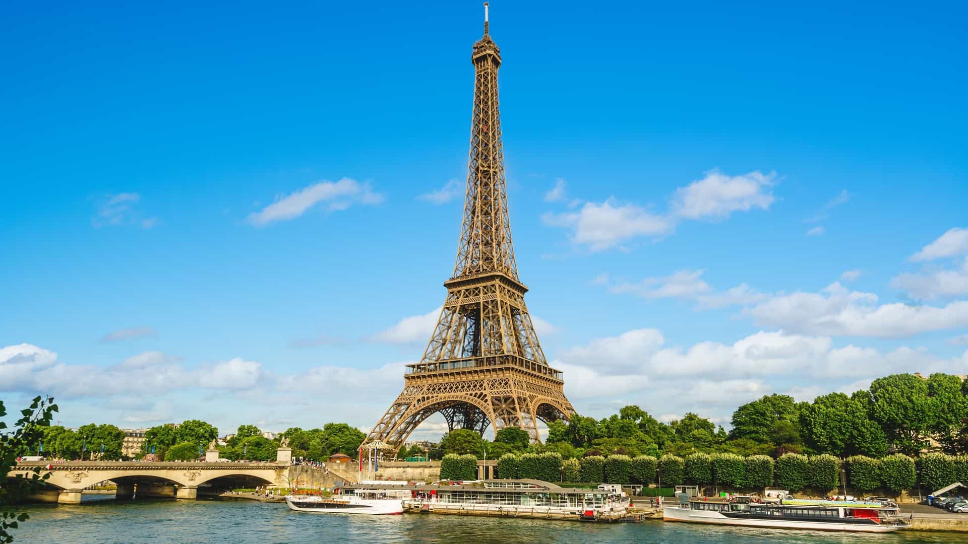 A classic cityscape view of the Eiffel Tower in Paris, France, as seen from or across the Seine River. The famous wrought-iron lattice tower rises prominently from the Left Bank, with the river, bridges, and surrounding urban skyline (including buildings like Les Invalides) often visible in the foreground or background, capturing the iconic and romantic atmosphere of the French capital.