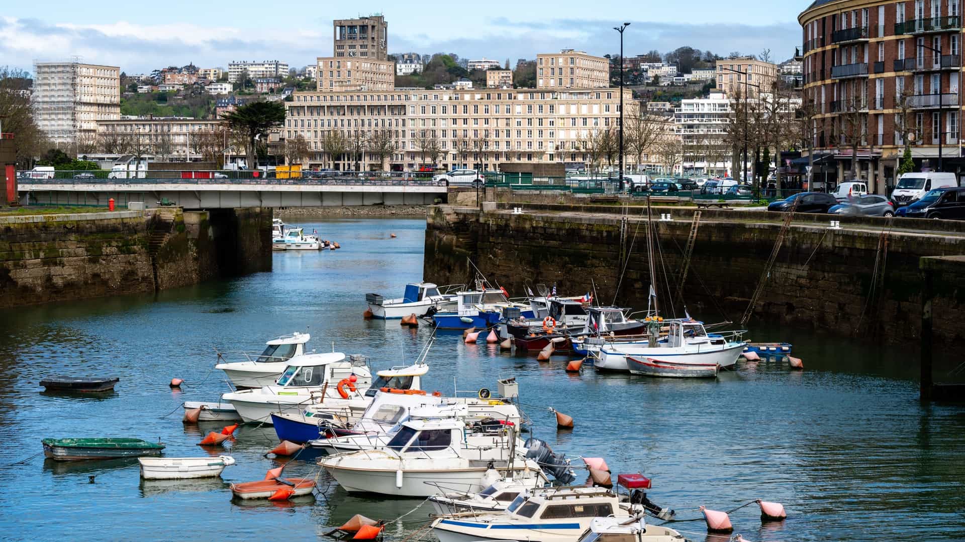 A vibrant view of the Port of Le Havre (Port de Plaisance) in Normandy, France. The foreground features a collection of pleasure boats and yachts docked in a basin or marina. In the background, the city's unique post-WWII modernist architecture—part of the UNESCO World Heritage site—rises above the quayside. The image highlights Le Havre's identity as a major maritime city, blending its role as a working port with a recreational waterfront.