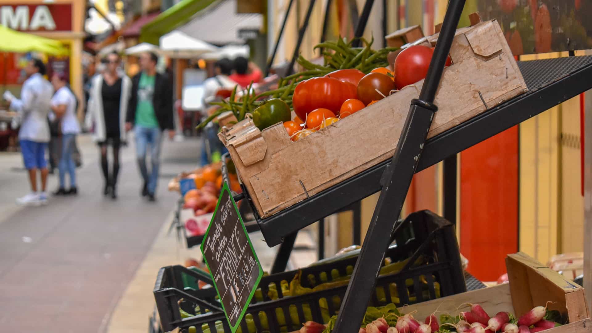 Shop at Le Havre's Local Markets. A vibrant scene inside a covered market in Le Havre, France, showing a stall heavily laden with a colorful variety of fresh fruits and vegetables. The produce is neatly arranged in baskets and crates, highlighting the local selection available at the market.