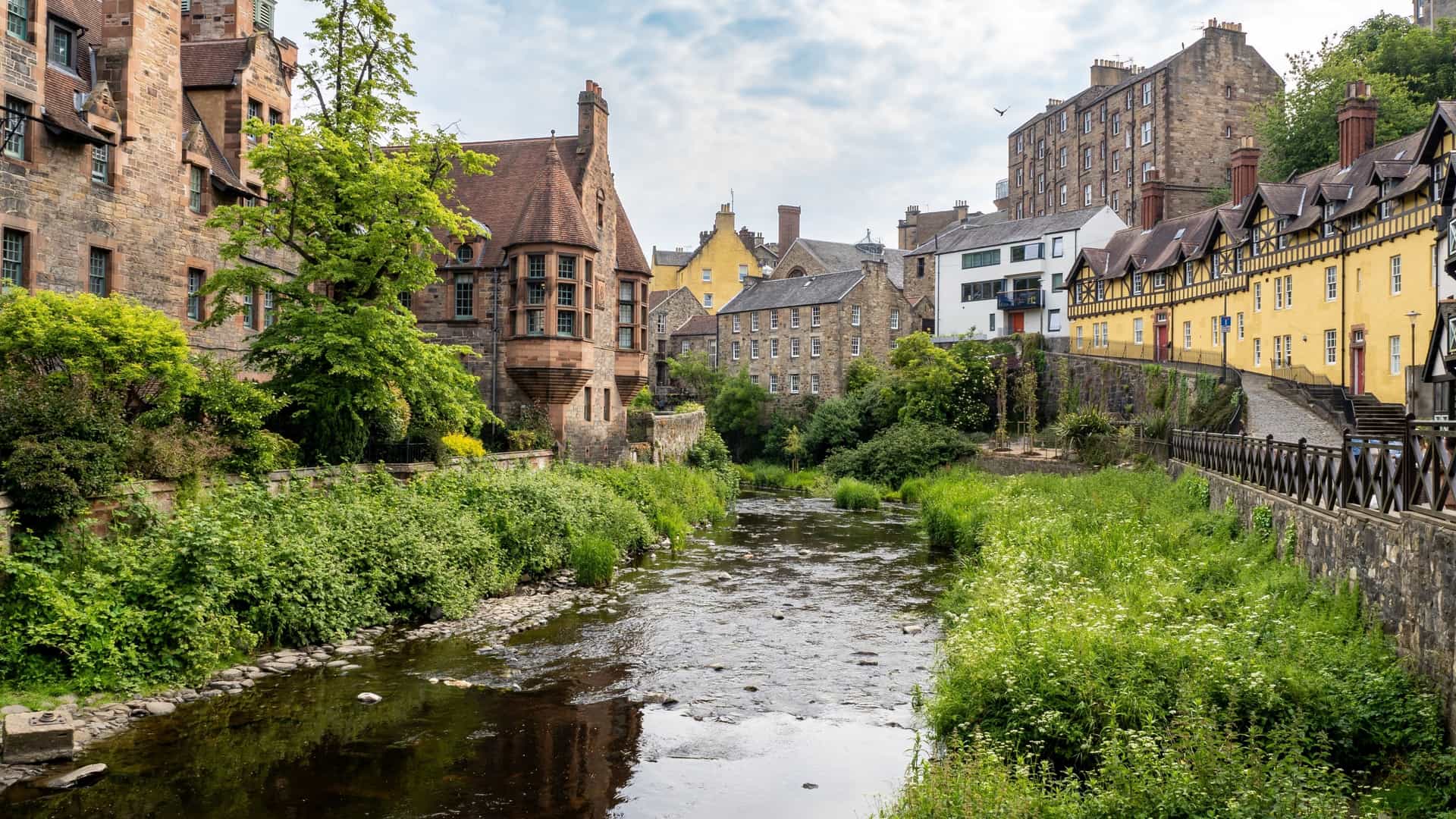 A scenic view of the Water of Leith Walkway in Edinburgh, Scotland. The path runs alongside the Water of Leith river, which is surrounded by lush green trees. The image captures the tranquil atmosphere of the walkway, a popular path for walking and cycling.
