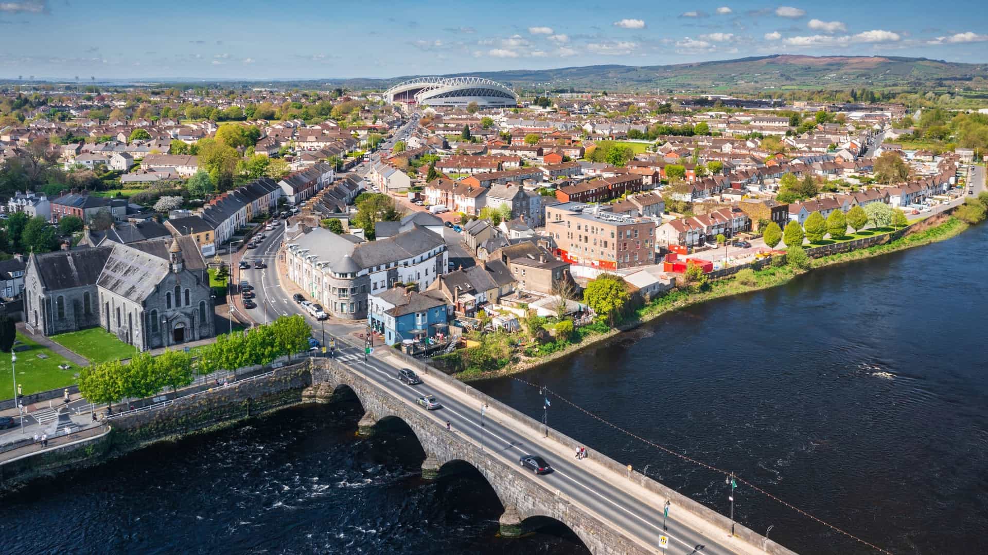 An aerial view of Limerick City, Ireland, featuring the Sarsfield Bridge crossing the River Shannon. The prominent Georgian building on the riverbank near the bridge is the historic Hunt Museum (the former Custom House), set against a backdrop of residential areas and the surrounding hills.