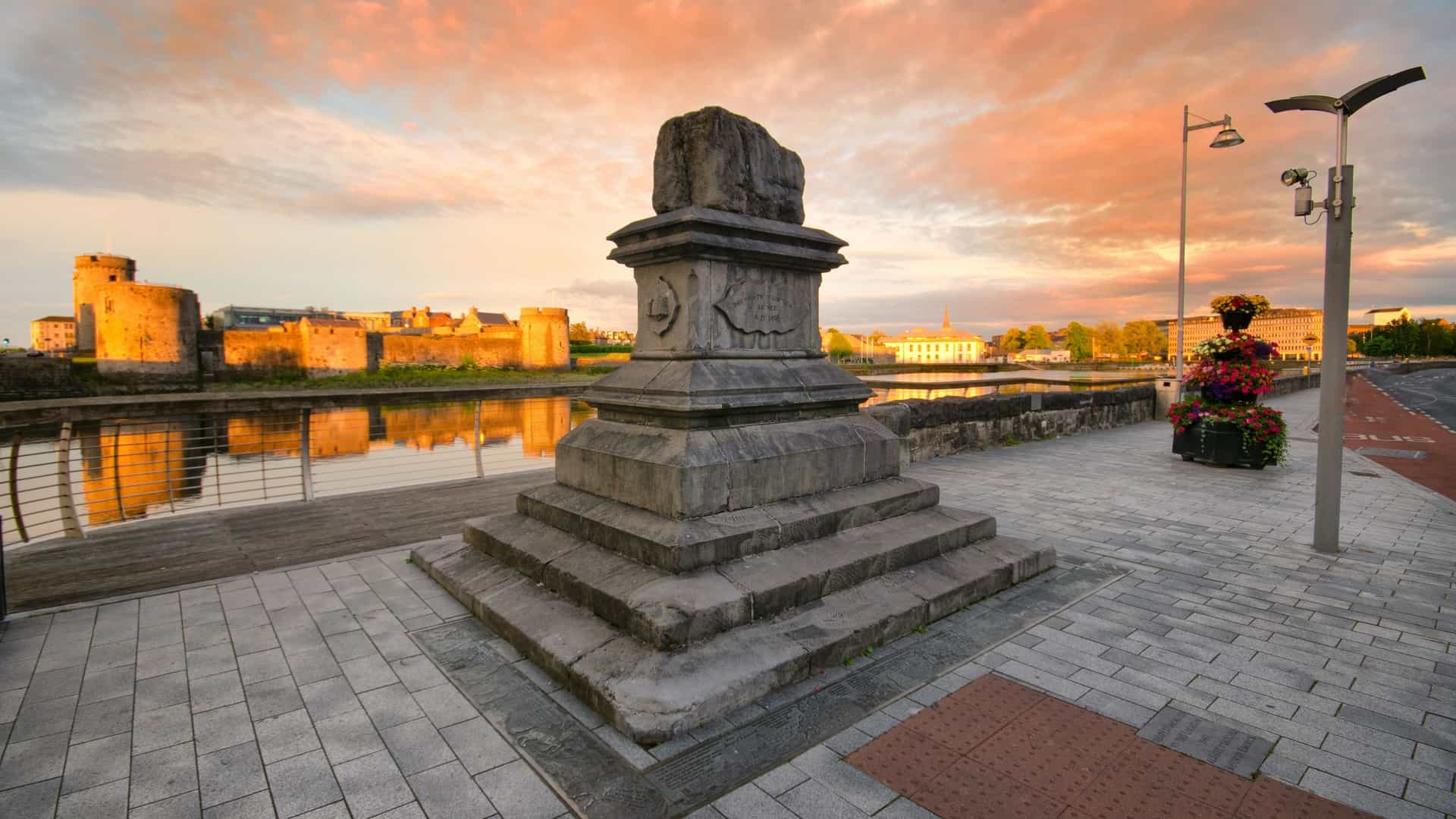 A panoramic view of Limerick City, Ireland, at sunset. The historic Treaty Stone monument on a plinth is prominent in the foreground on the riverbank, with the River Shannon flowing behind it, and the imposing medieval King John's Castle visible across the water on King's Island.