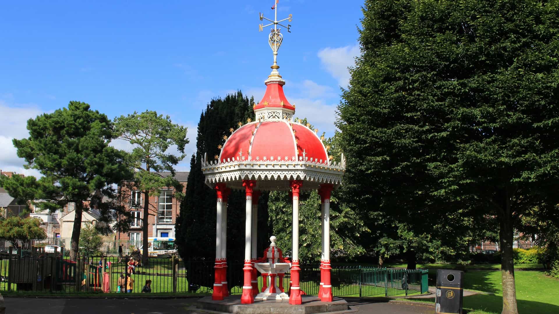 A photo of the Richard Russell Fountain, a historic, ornate, red-and-white painted cast-iron drinking fountain, located within the green, tree-lined People's Park in Limerick City, Ireland.