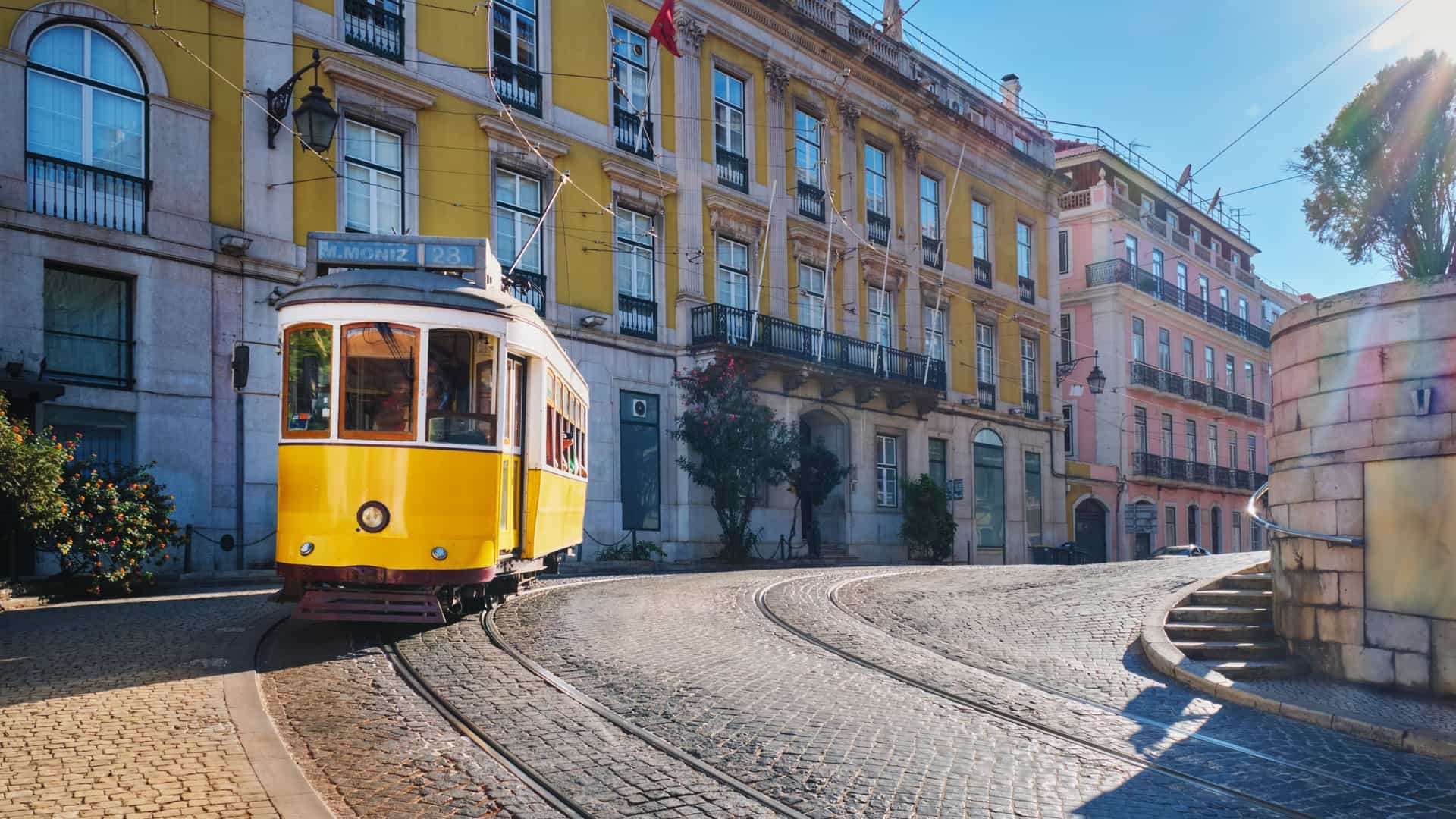 A famous vintage yellow Tram 28 navigates a narrow, cobblestone street in the Alfama district of Lisbon, Portugal. The historic tram, a classic symbol of the city, is seen moving past traditional buildings.