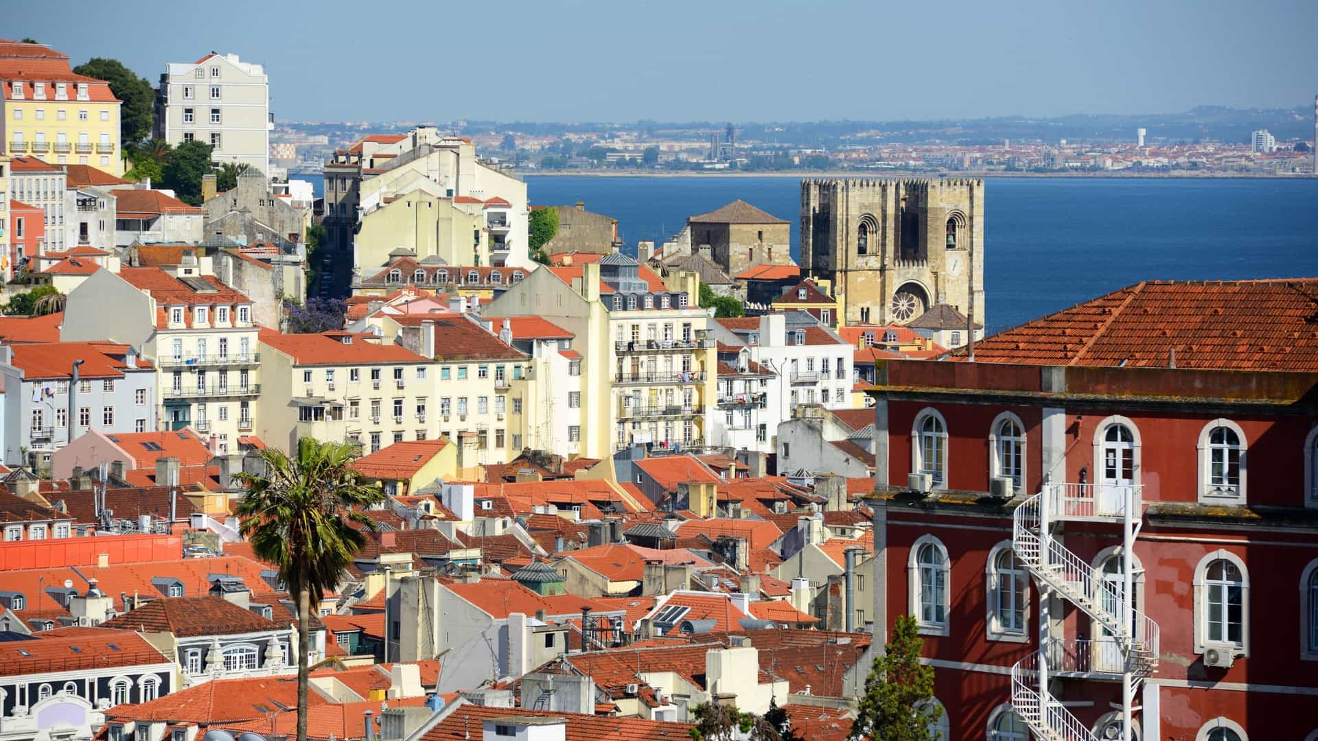 A high angle view of the Lisbon Cathedral (Sé de Lisboa), the oldest church in the city, with the Tagus River visible in the background. The cathedral's fortress like Romanesque facade with two bell towers is prominently featured, set against the backdrop of Lisbon's historic rooftops.
