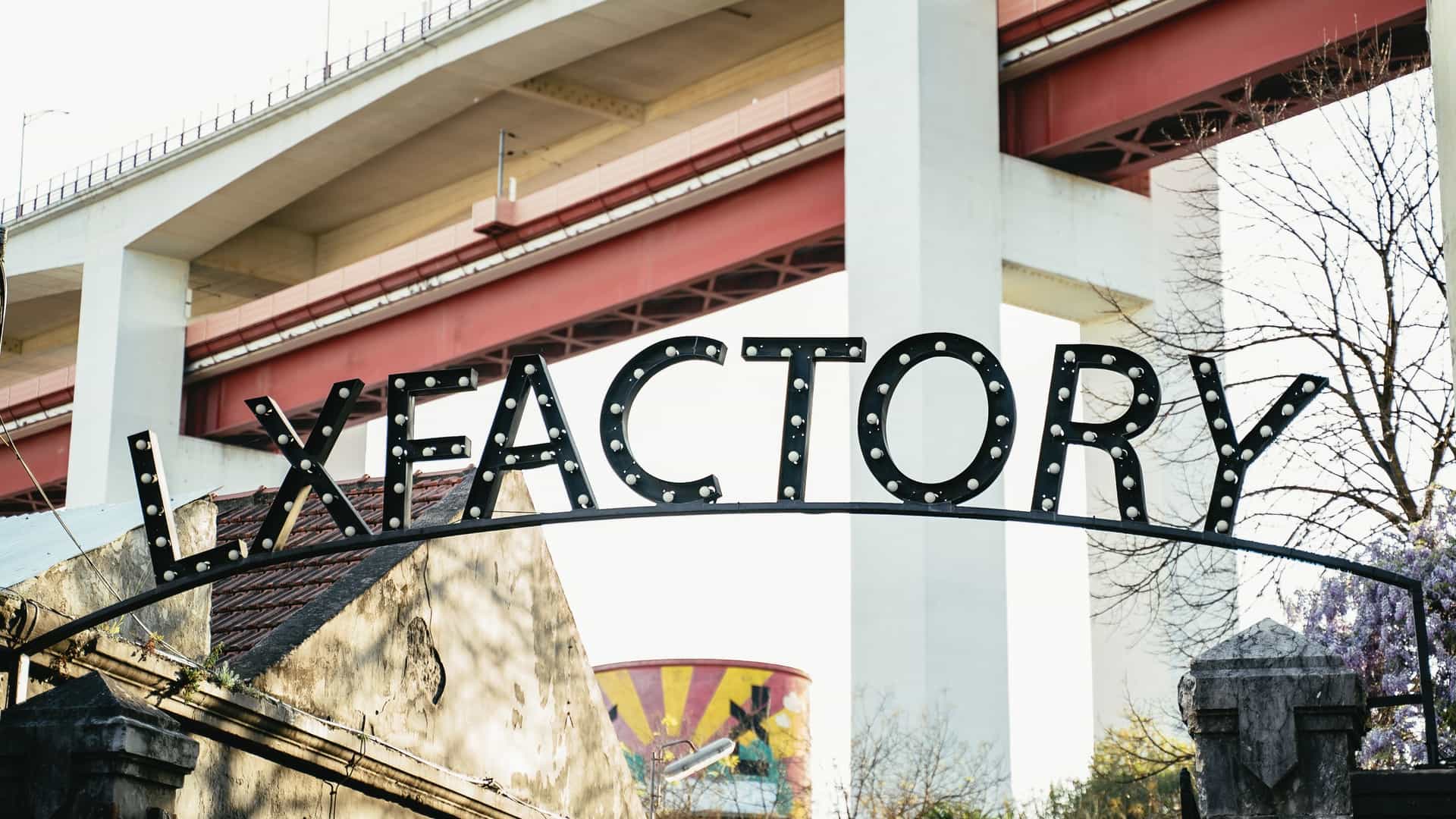 A close-up of a weathered, industrial-style sign reading "LX Factory" in a converted industrial complex in Lisbon, Portugal, with a red architectural detail visible against a building facade.