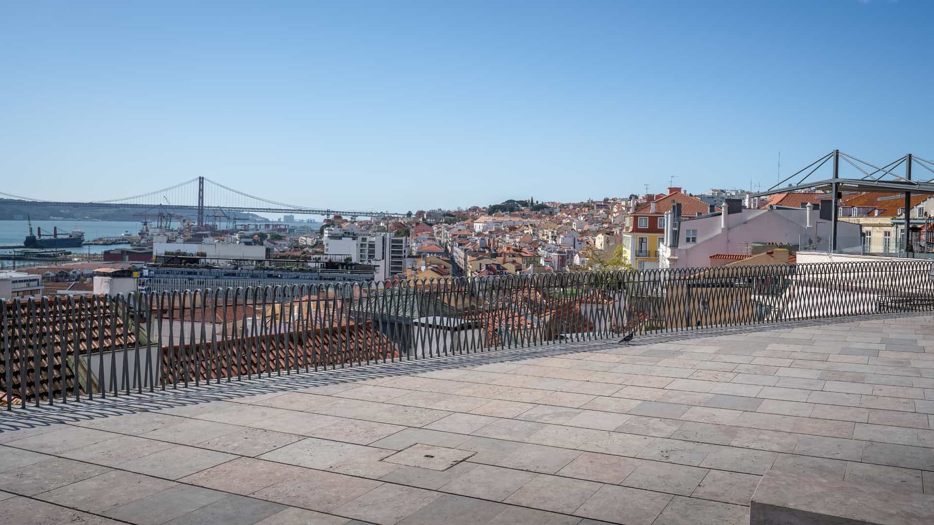 A panoramic view from the Miradouro de Santa Catarina viewpoint in Lisbon, Portugal. The image shows the red rooftops of the city leading down to the Tagus River, with the suspension bridge, Ponte 25 de Abril, and the Cristo Rei statue visible in the distance. The foreground features a stone terrace with a statue of the mythical sea monster, Adamastor.