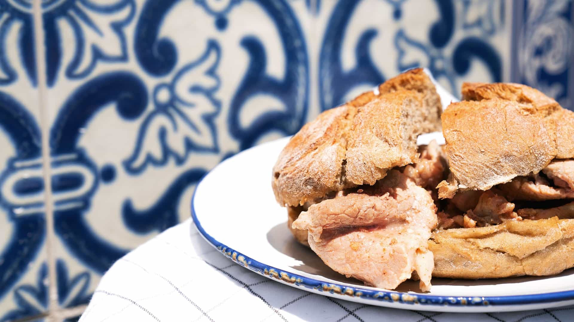 A close up shot of a traditional Portuguese Bifana sandwich. The sandwich features thin slices of marinated and cooked pork piled inside a soft, crusty Portuguese bread roll. The pork appears tender and moist, with a rich, savory color.